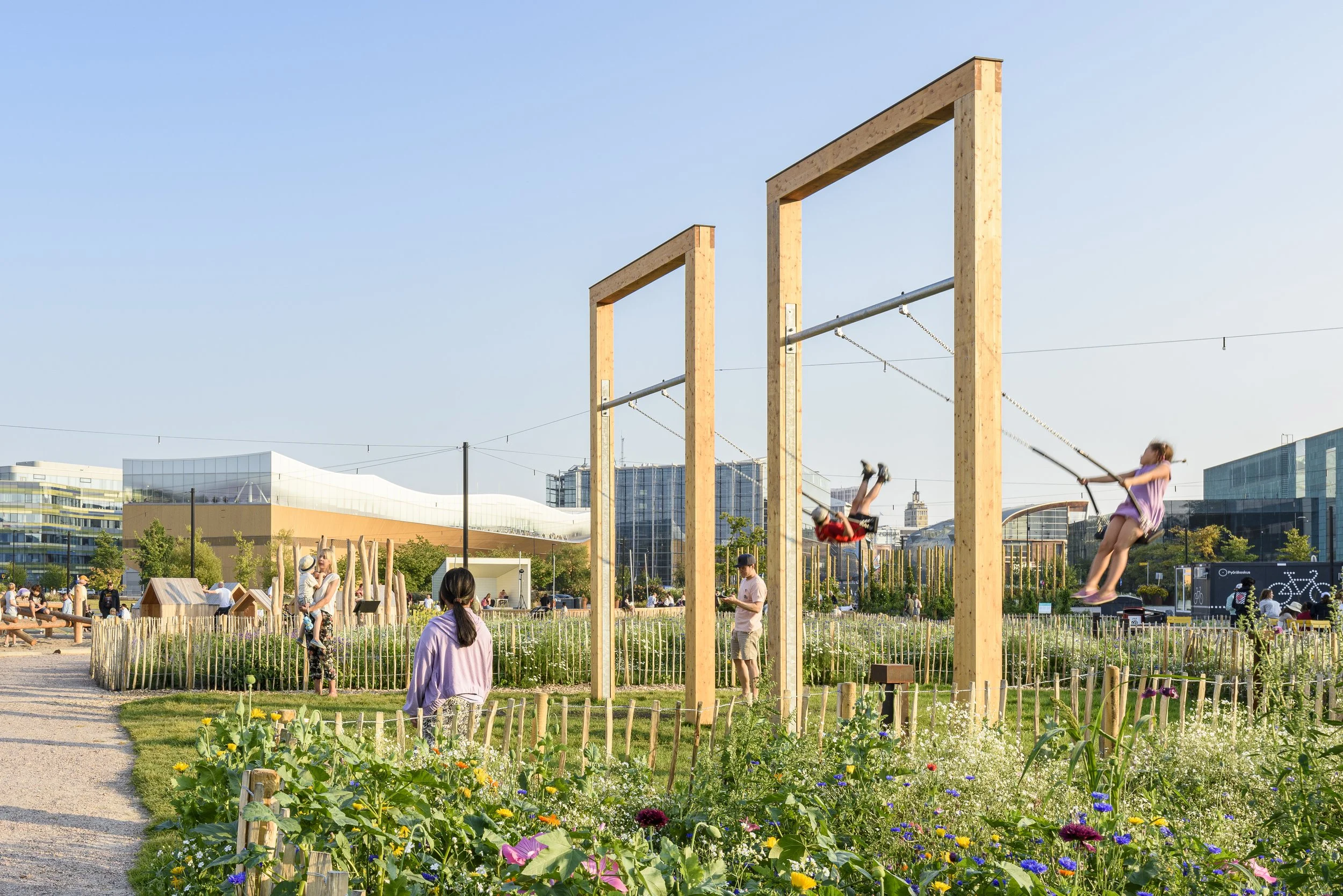 Helsinki new Park.
Children playing on swings in a park with modern buildings and a garden with flowers in the background.