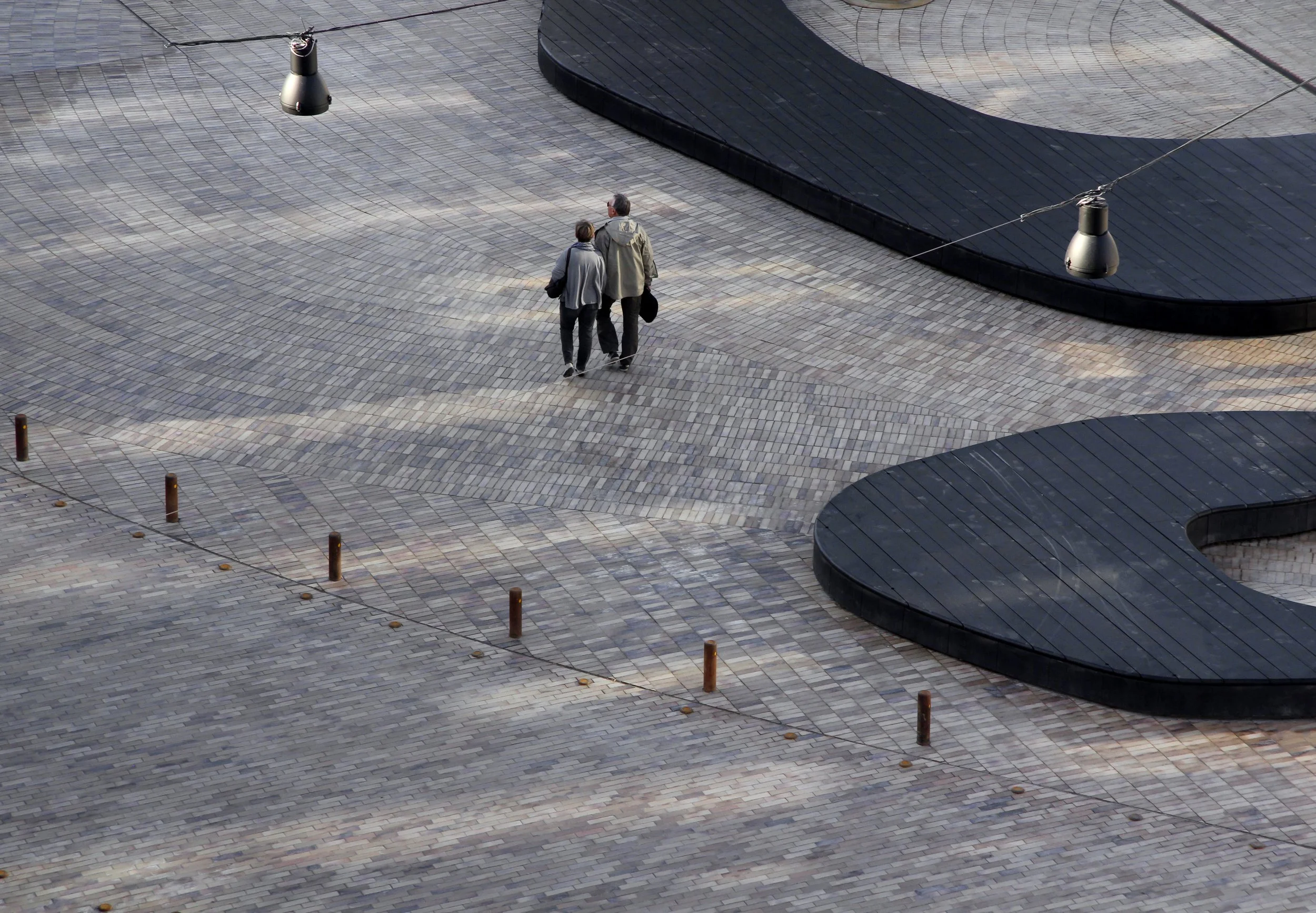 Two people walking on a large paved outdoor area with a pattern of gray tiles and black raised sections. There are streetlights and safety posts, with cables connecting the lights.