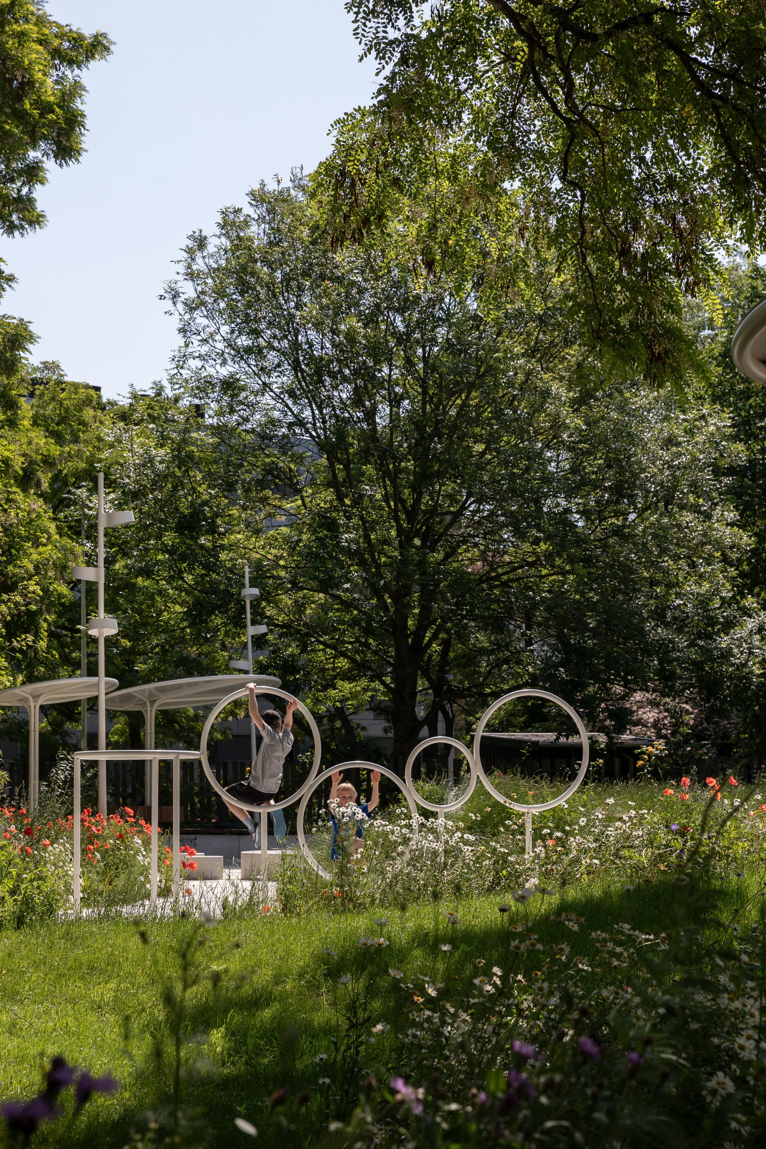 Nørrebro landscape. Urban renewal.
Children playing on modern circular playground equipment in a lush green park with trees and colorful flowers.