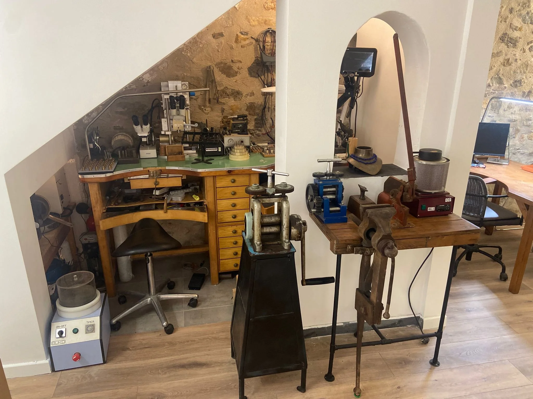Atelier de bijouterie avec divers outils et machines, incluant un tour de bijoutier, une table de travail, et un établi avec microscopes et autres instruments.