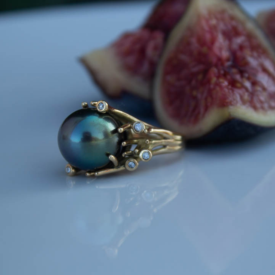 Bague avec une perle noire et des diamants, accompagnée d'un figue fraîche, sur une surface lisse.