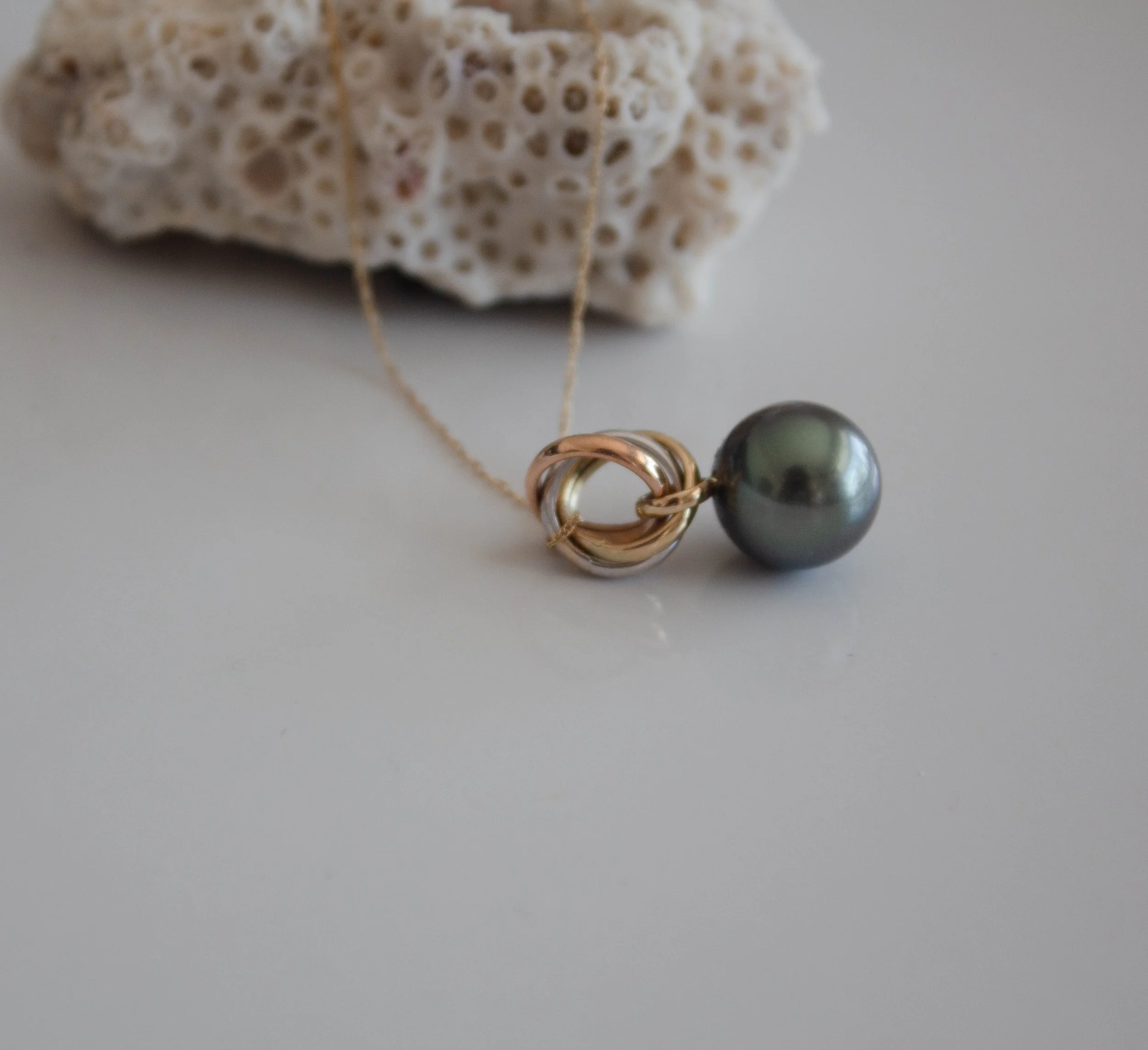 Pendentif en or rose avec une perle noire et un nœud en or, posé sur une surface blanche avec une pièce de corail en arrière-plan.