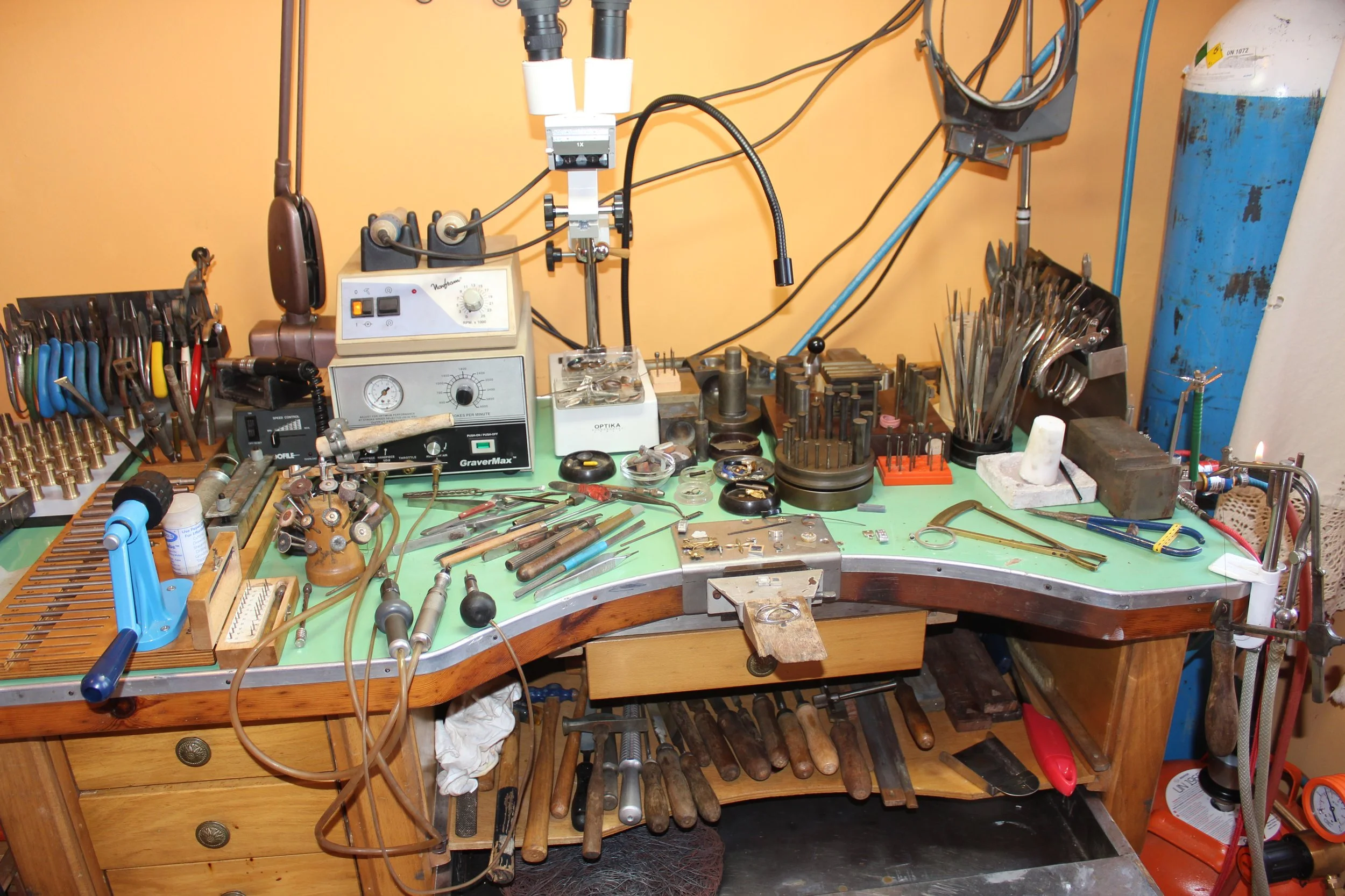 Un établi de bijoutier avec divers outils de bijoux, comme des pinces, des marteaux, une loupe, des outils de gravure, et d'autres instruments, sur un plan de travail vert, avec un fond beige.