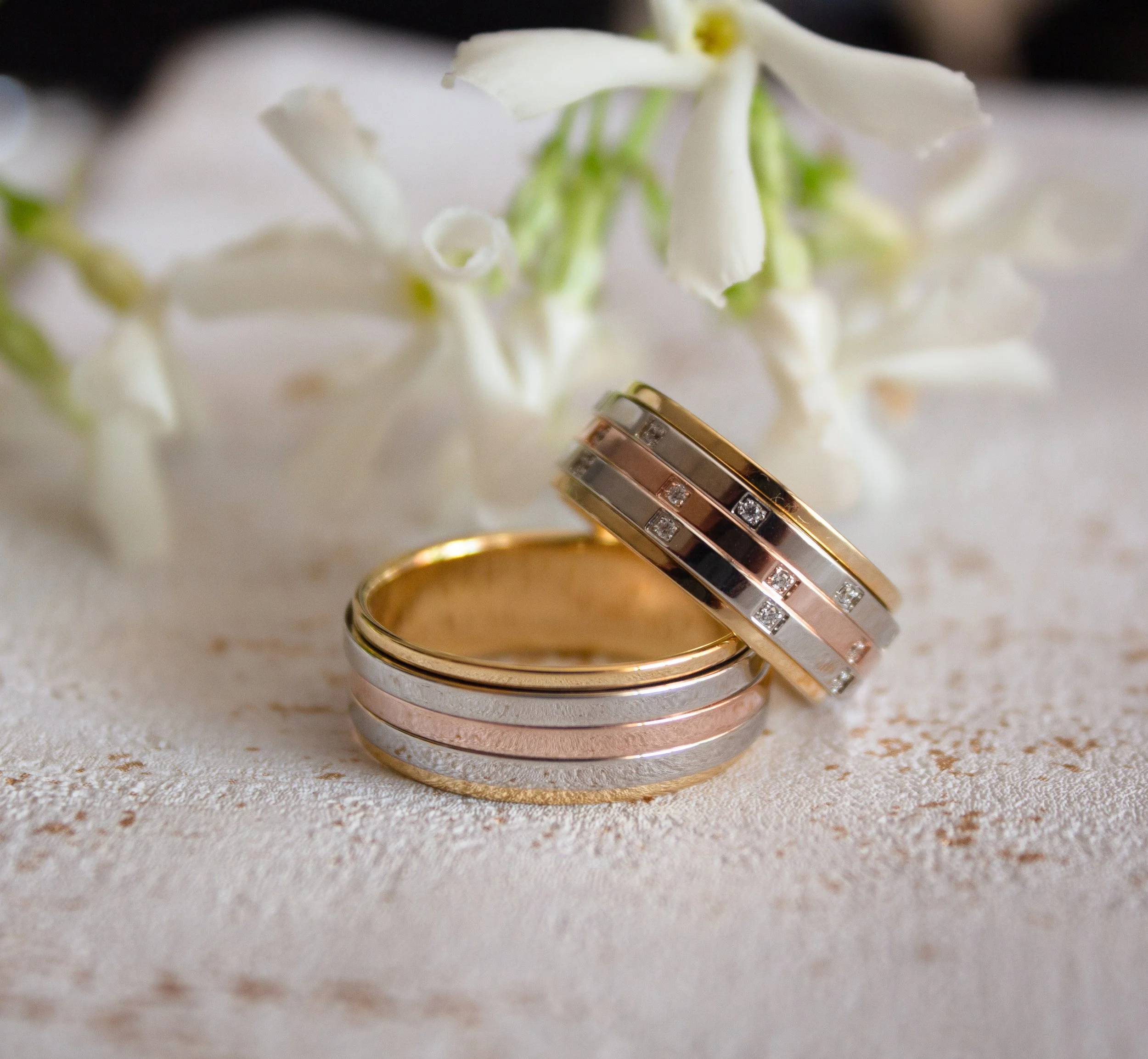 Deux alliances de mariage en métal or, argent et rose avec des diamants, posées sur une surface claire avec des fleurs blanches en arrière-plan.