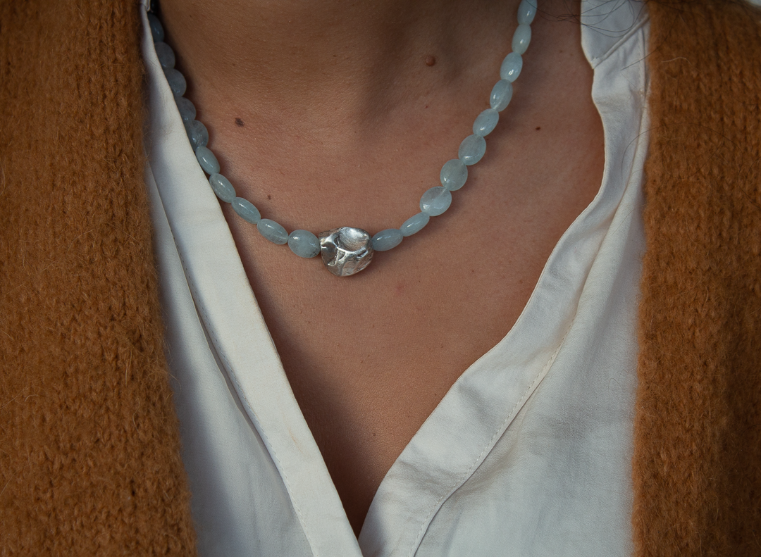 Collier de perles bleues avec un pendentif en forme de coquillage en argent, porté avec une chemise blanche et un pull marron.