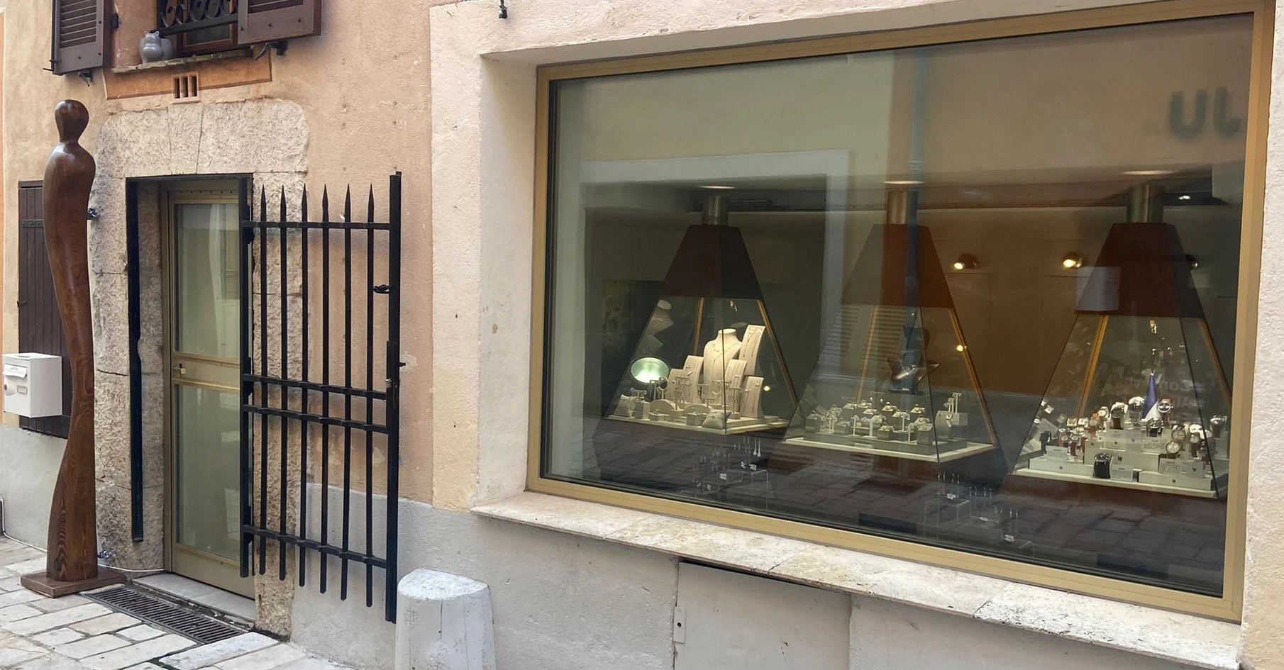 Vitrine d'une bijouterie avec montres et bijoux exposés à l'intérieur, vue depuis la rue, bâtiment en pierre avec une sculpture en bois à gauche.