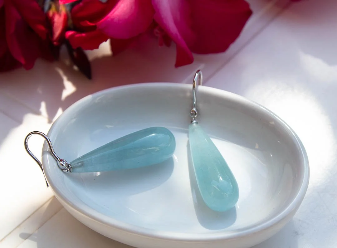 Boucles d'oreilles en pierre aigue-marine dans un bol blanc, fond avec fleurs roses, lumière naturelle.