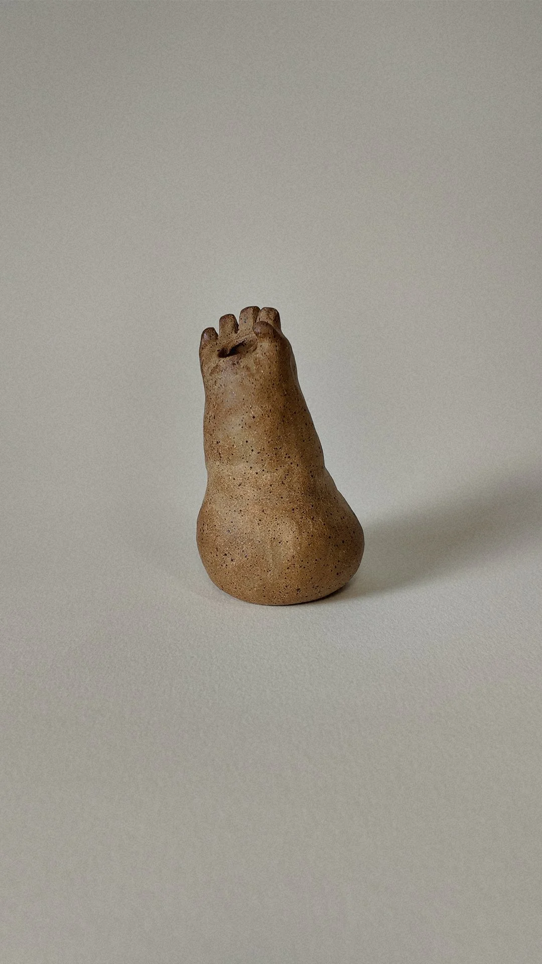 Small sculpture resembling a human foot, made of textured material with a natural, earthy color, placed on a plain, light-colored surface.