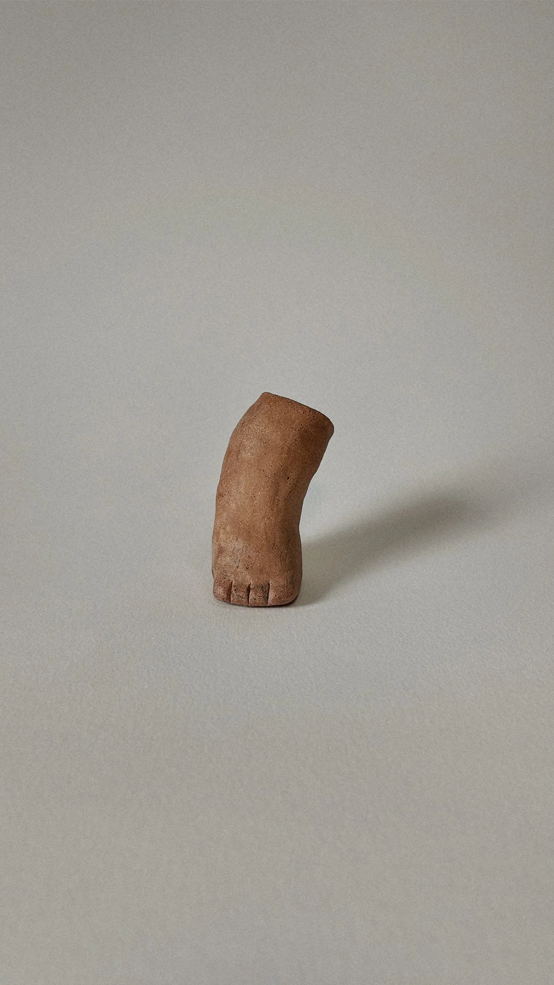 A small, figurative sculpture of a foot made from a clay-like material, standing upright on a plain surface against a simple background.