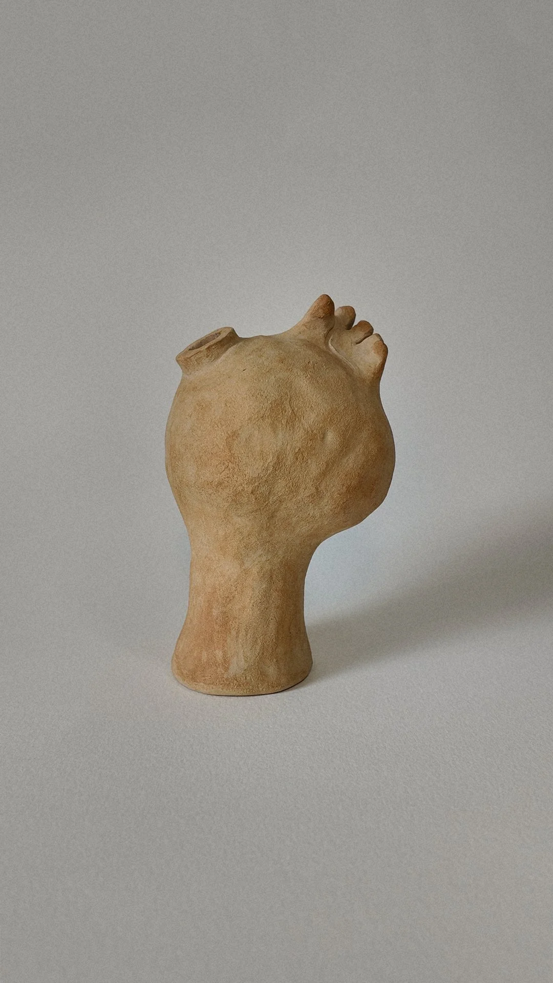 A sculpture of a human head with a face but no distinct features, mounted on a short neck, made of a textured, beige material, set against a plain gray background.