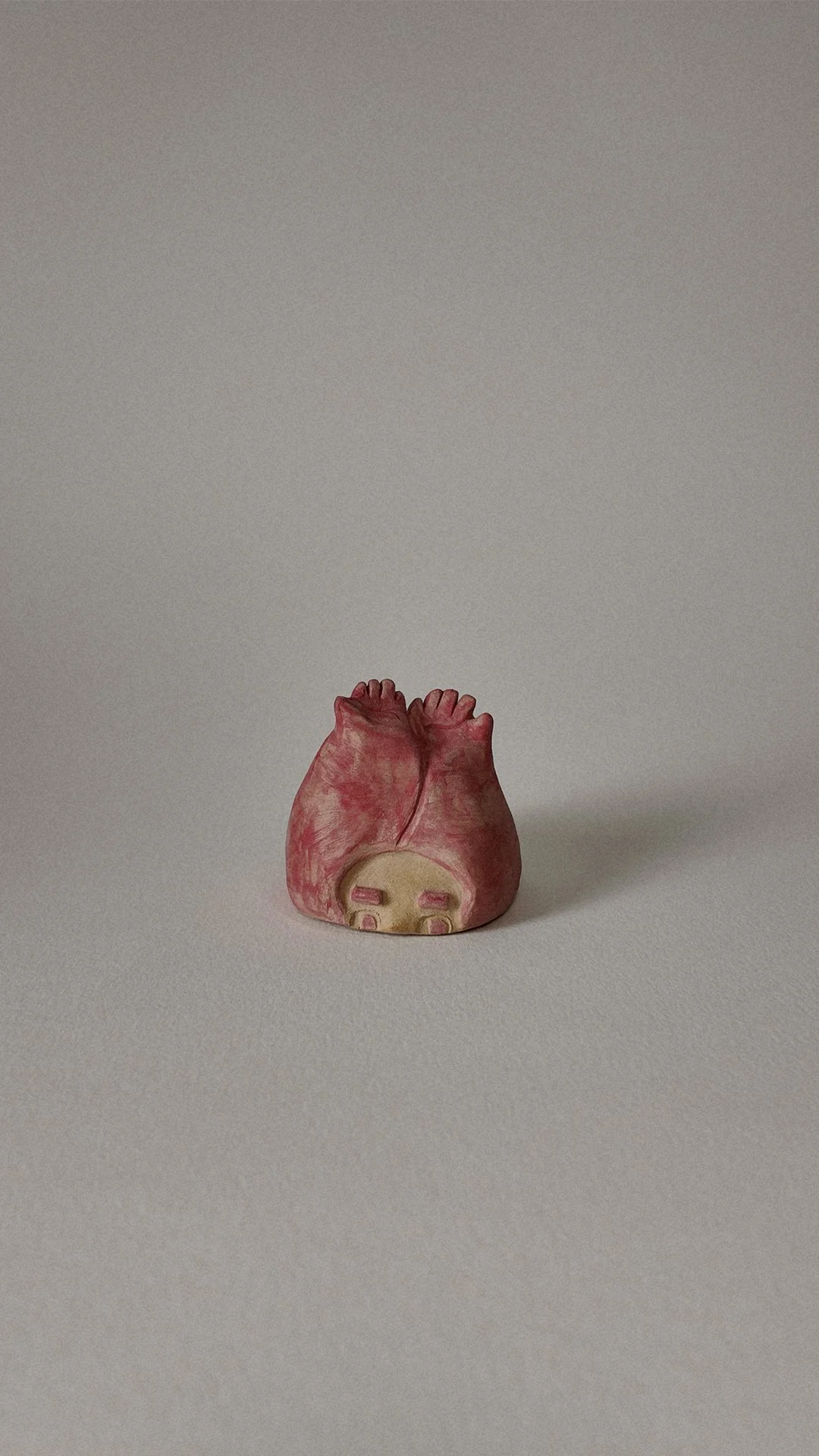 Small pink ceramic heart-shaped object with facial features on a plain gray background.