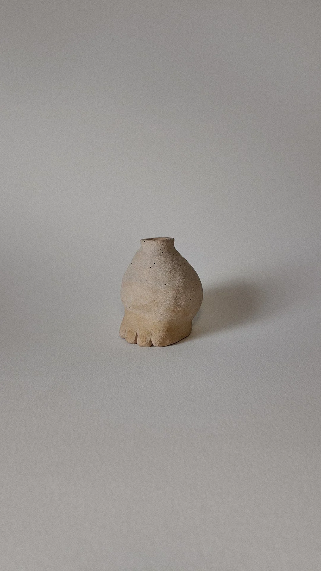 Small beige ceramic vessel with a round body and a short, narrow opening, featuring decorative indentations at the base, placed on a plain light-colored surface.
