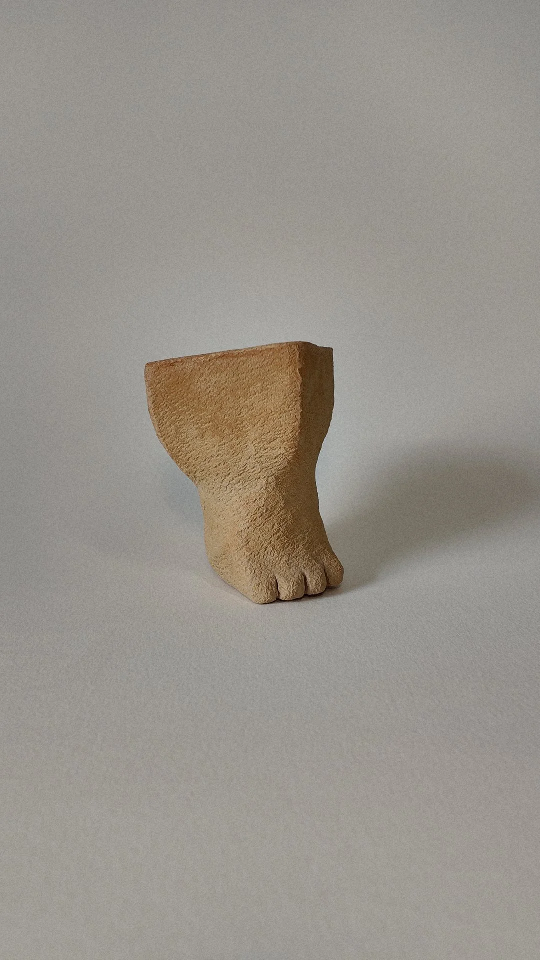 A small sculpture of a foot with toes, made of a textured material, positioned on a plain surface with a shadow to its right.