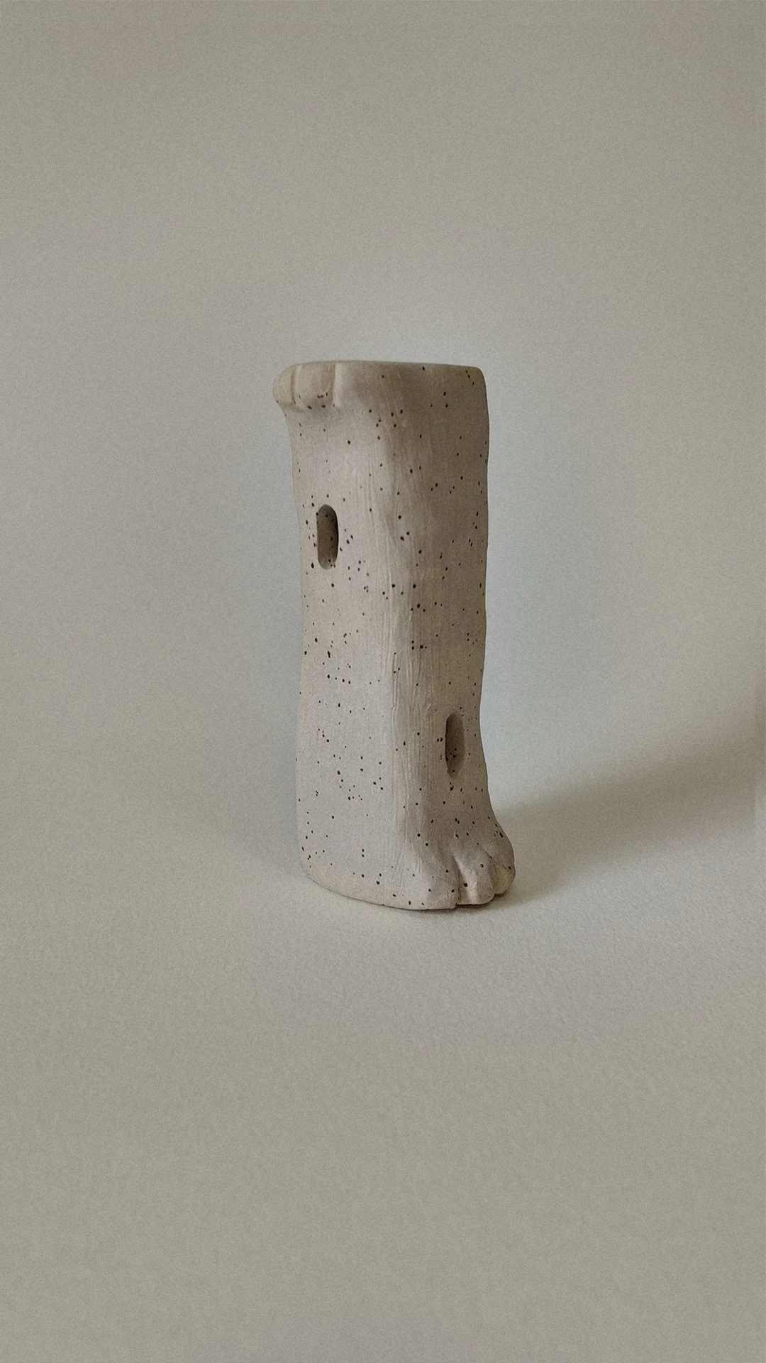 A minimalist beige ceramic sculpture with small holes, resembling a face or abstract figure, against a neutral background.