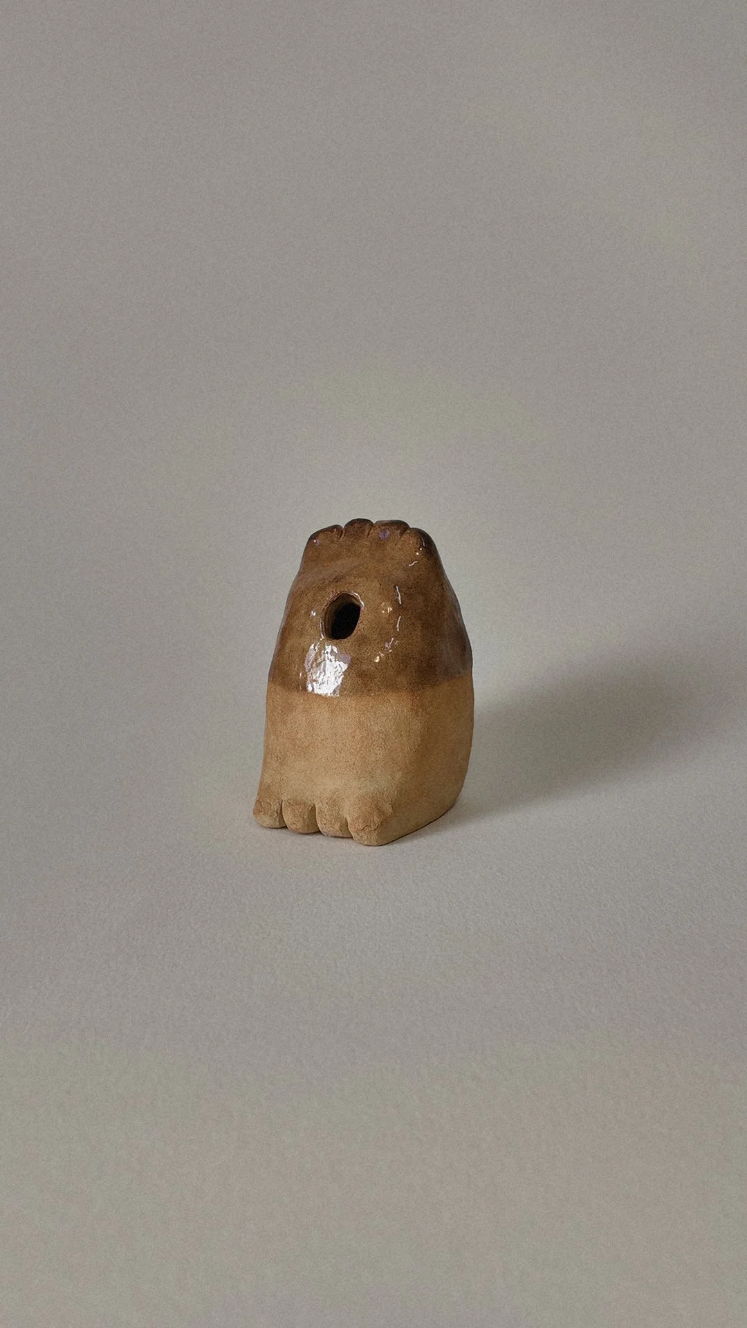 Small ceramic figurine resembling a creature with a hole for an eye and textured feet, glazed in shades of brown and beige.