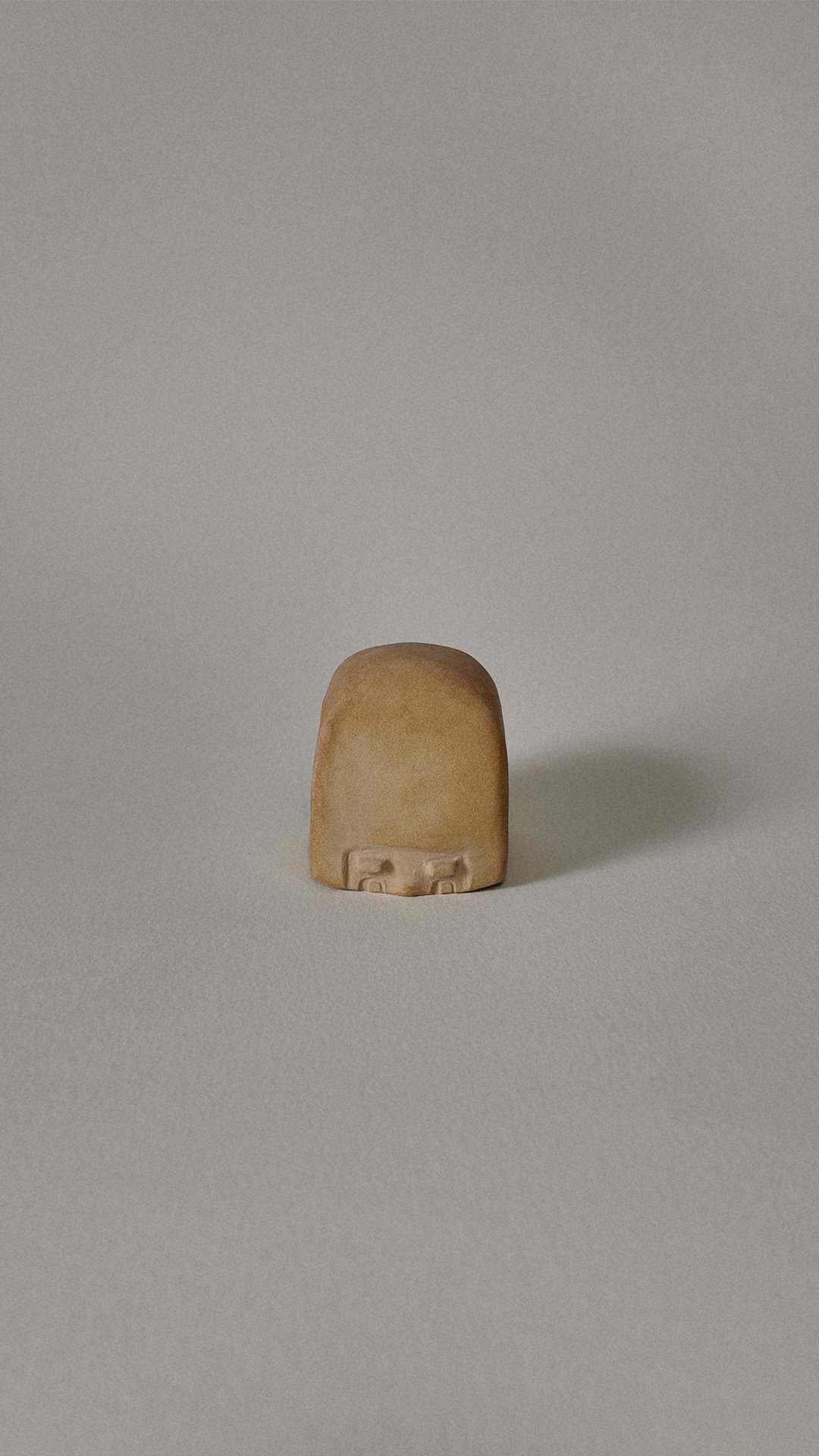 A small, beige, rounded ceramic figure resembling a stylized human face with minimal features, set against a plain, light-colored background.