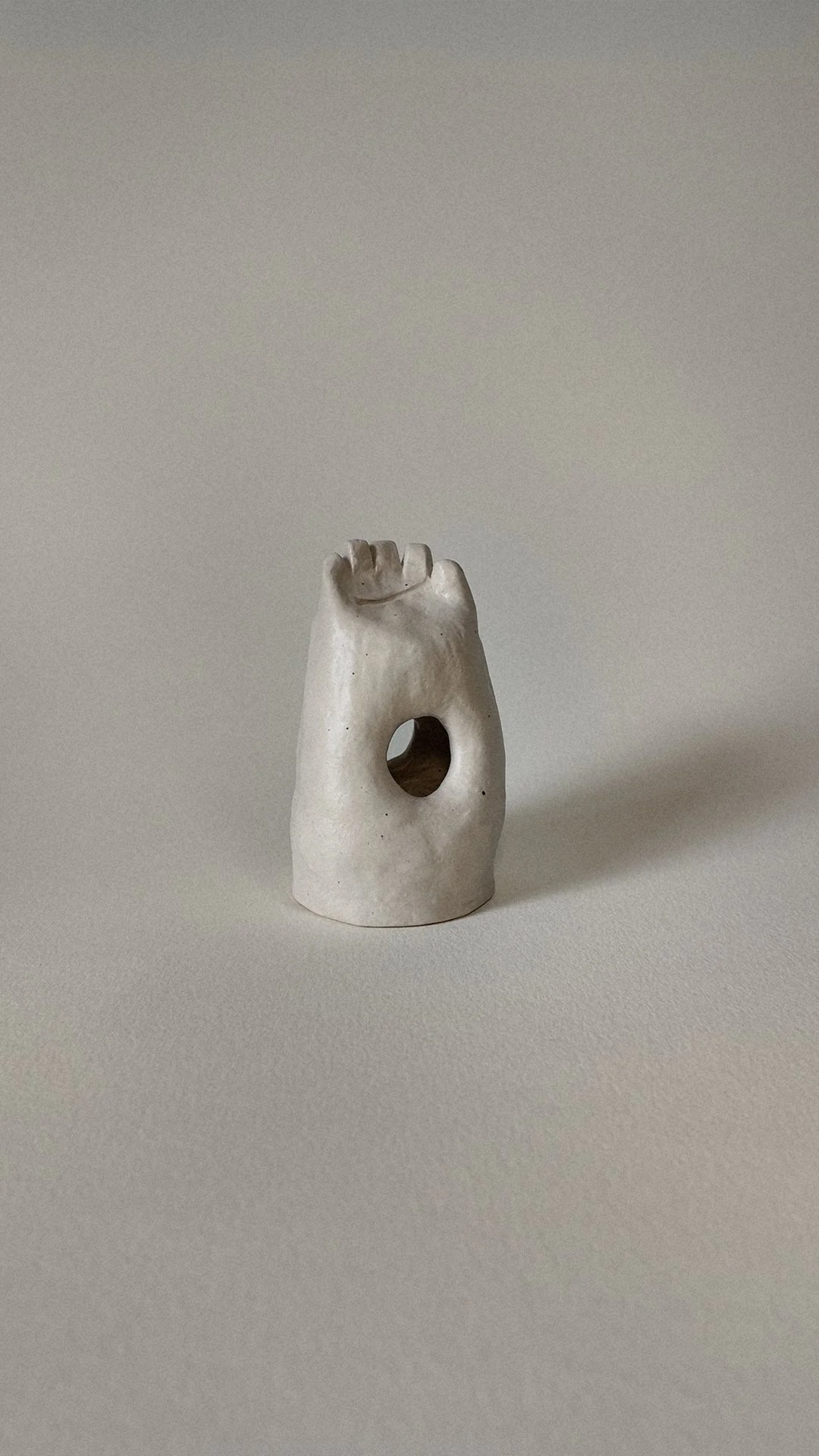 A small white ceramic sculpture resembling a human figure with a hole in the middle and a hand on top.