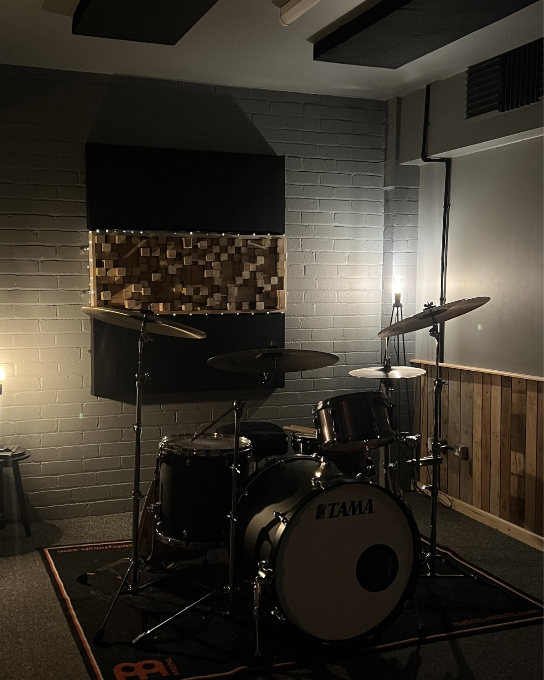 A black Tama drum set with cymbals on stands in a music studio corner. Behind it, there are acoustic foam panels and wooden soundproofing on the wall, with a dimly lit industrial-style lamp.
