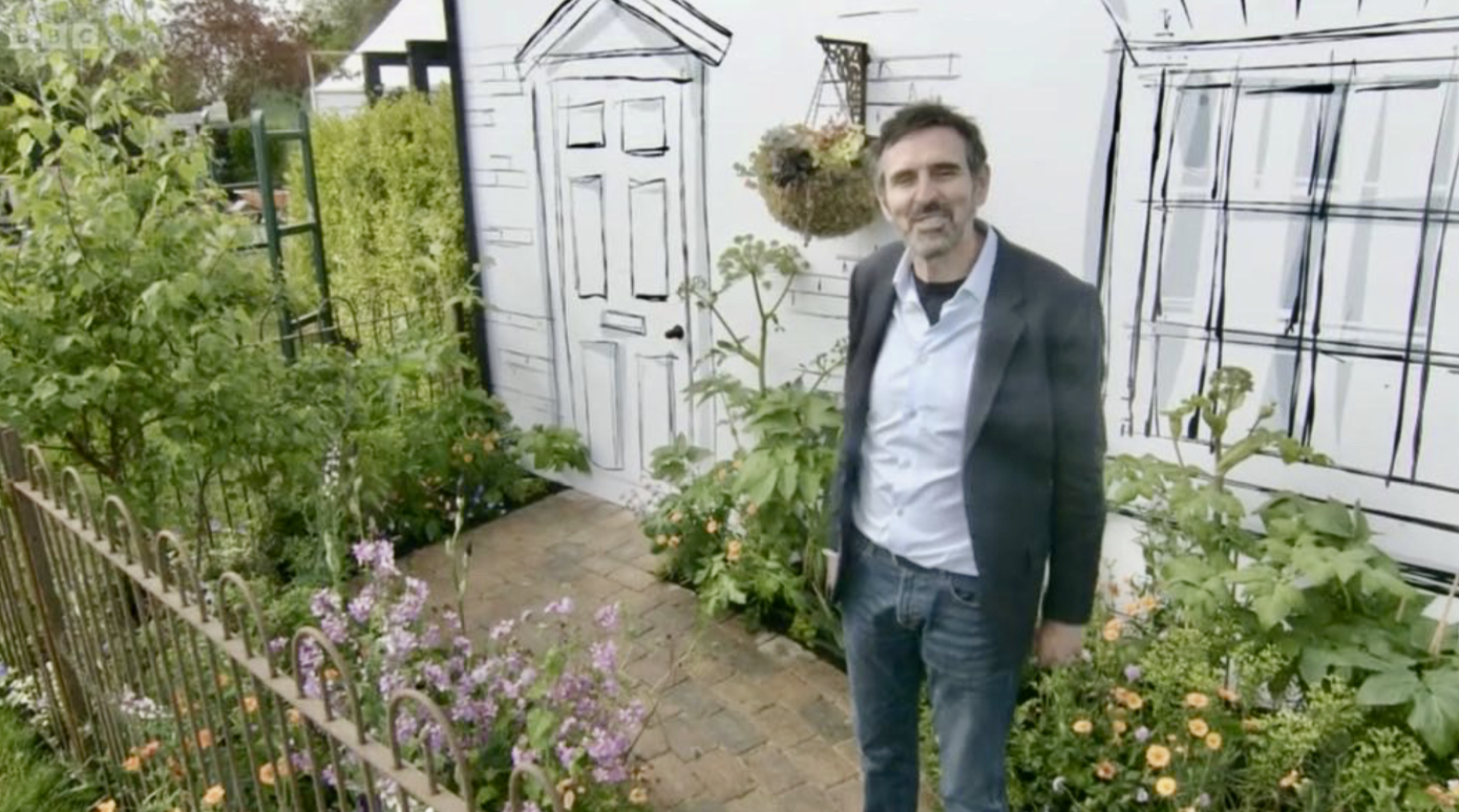 Adam Frost at RHS Malvern, next to a white wall with a black window outline and door illustration, and a hanging flower basket.