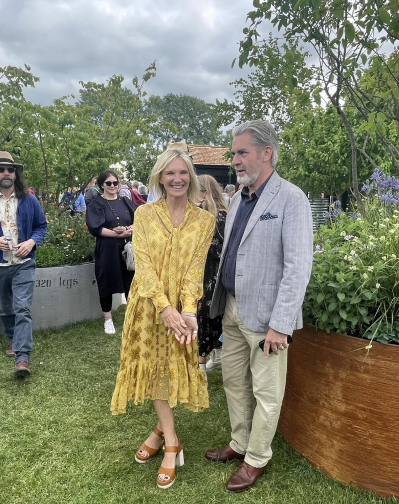 Jo Whiley and Mark Glynne-Jones at RHS Malvern flower show. The background features green trees, cloudy sky, and other attendees.