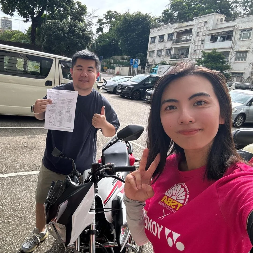 A woman in a pink cycling jersey taking a selfie with a man in a black shirt holding a printed document and giving a thumbs-up in a parking lot, with motorcycles and cars in the background.