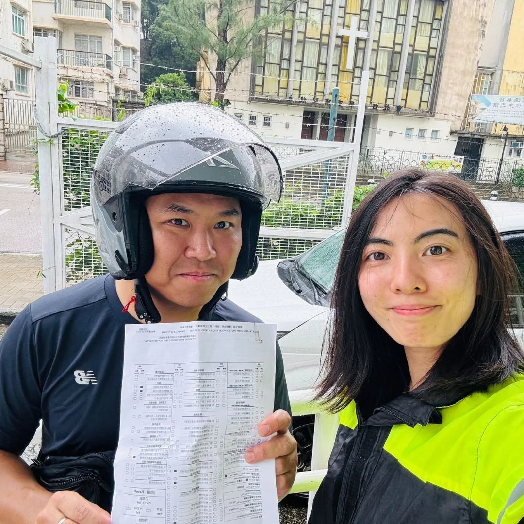 A woman and a man, both smiling, are taking a selfie outdoors on a rainy day, with the man wearing a black helmet and holding a document, and the woman wearing a high-visibility yellow jacket.