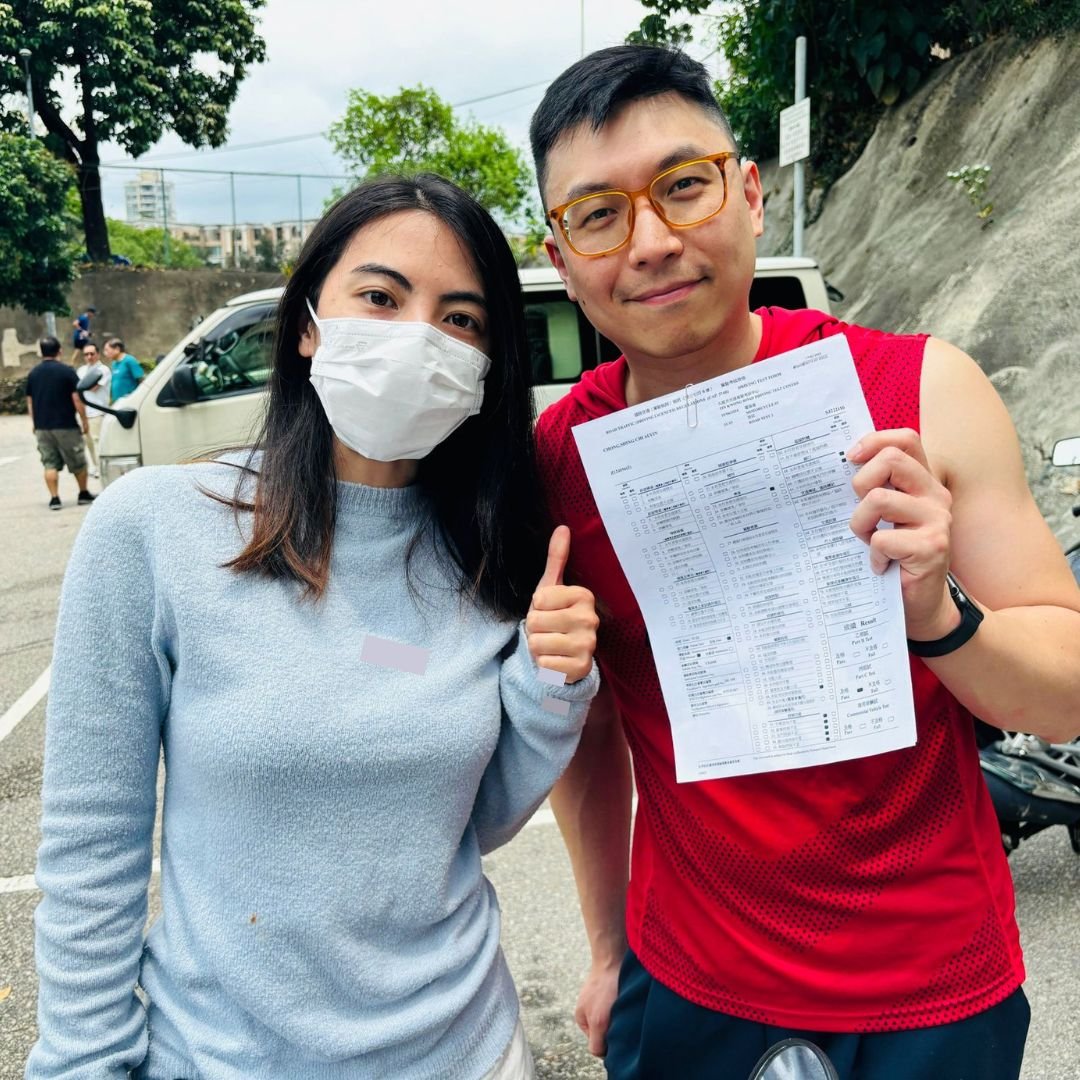 A man and woman taking a selfie outdoors, the woman wearing a face mask and the man holding a paper. Both are smiling and standing near a white vehicle, with other people in the background.