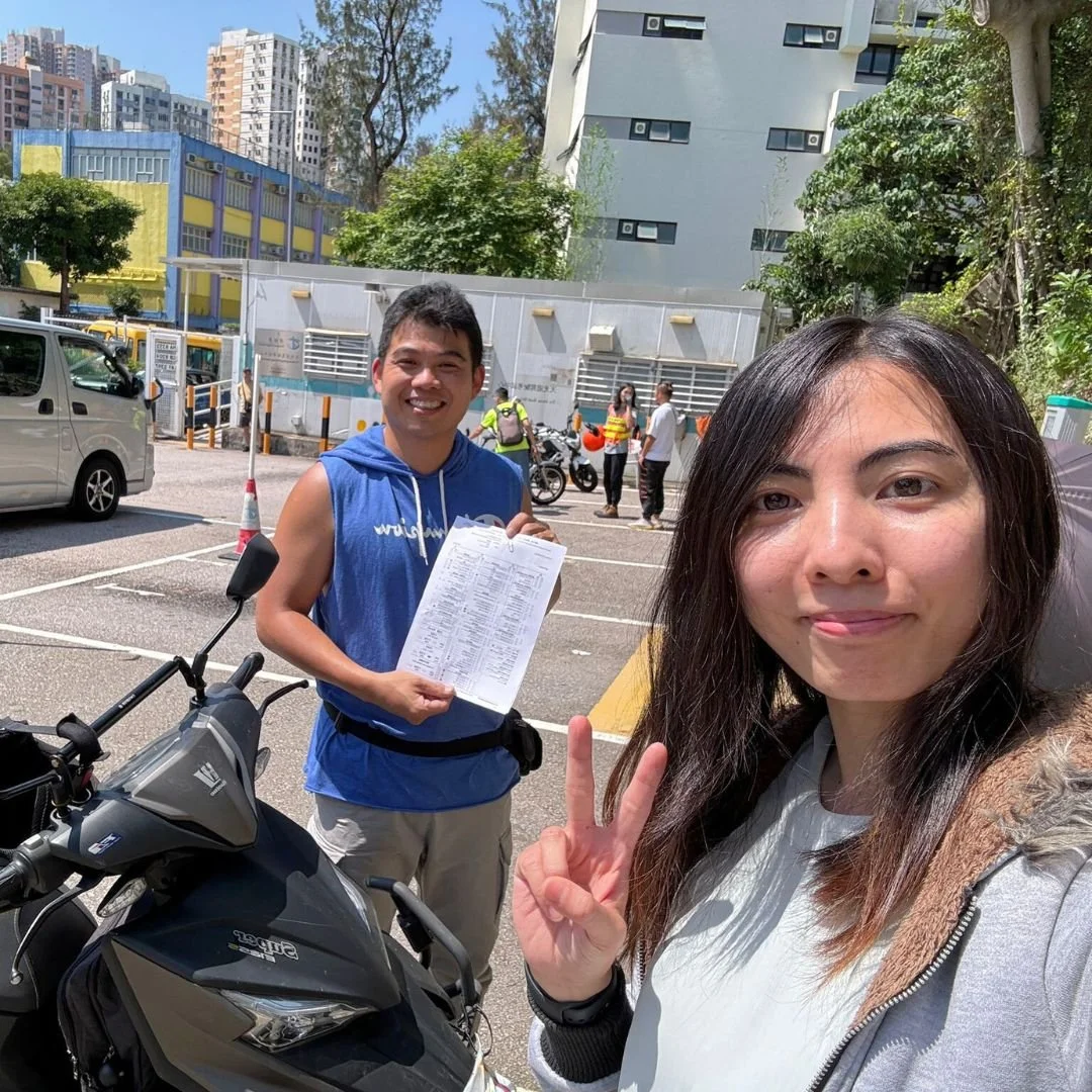 A woman taking a selfie with a man holding a clipboard in a parking lot on a sunny day. The woman is making a peace sign, and there is a motorcycle next to them. Several people and buildings are in the background.