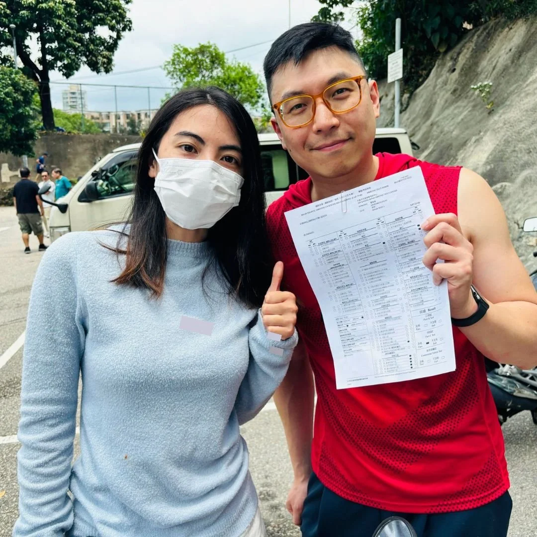 A man and woman outdoors. The woman wears a face mask and a light blue sweater, giving a thumbs-up. The man, wearing glasses, a red athletic shirt, and a wristwatch, holds a printed sheet of paper and smiles at the camera.