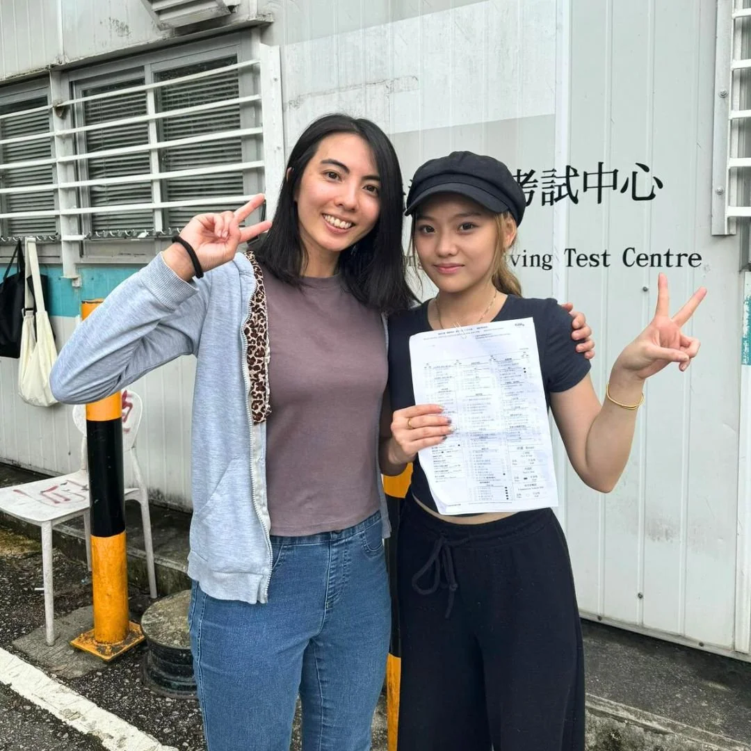 Two young women standing outside near a building with a sign that reads 'Driving Test Centre'. One woman is smiling and making a peace sign with her fingers, while the other woman is holding a paper and also making a peace sign. Both are casually dre