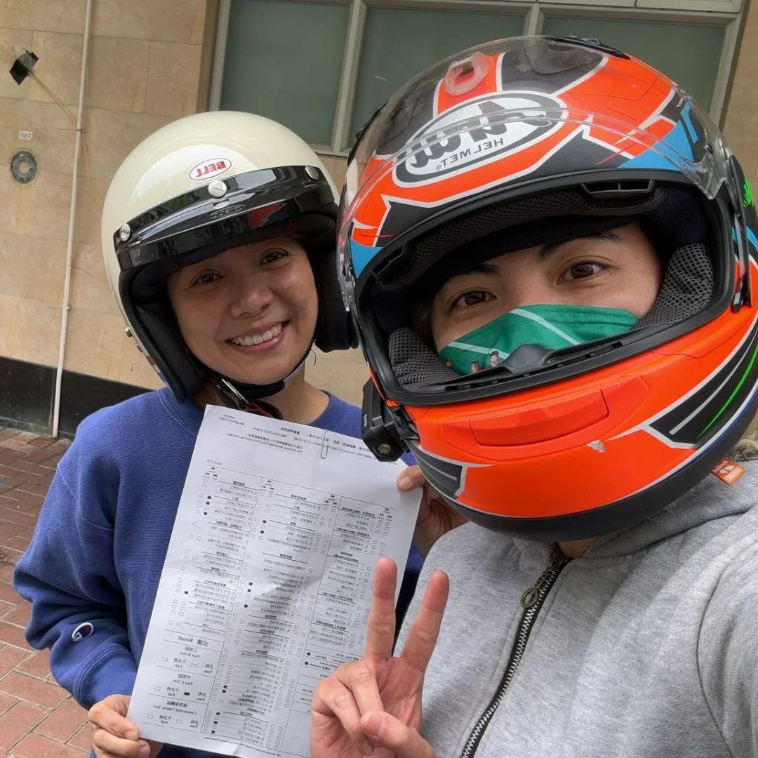 Two people wearing motorcycle helmets smiling for a selfie, one holding a test paper, and making a peace sign. They are outside near a beige building with windows.
