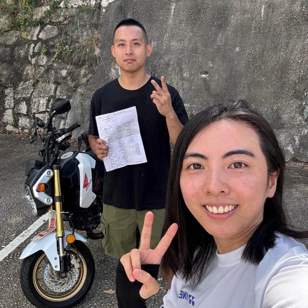 Two women taking a selfie outdoors with a motorcycle, a man holding a document and making a peace sign, and a motorcycle with a Honda logo in the background.