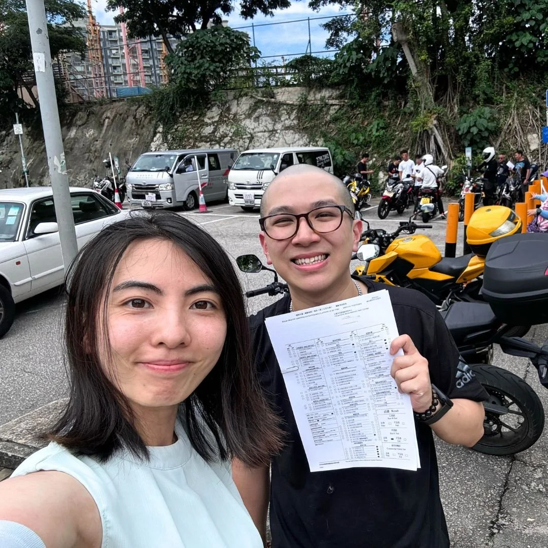 Two smiling people taking a selfie outdoors near a parking lot filled with cars and motorcycles. One person is holding a piece of paper, possibly a receipt or a document. Background shows a rocky hillside with trees and several parked vehicles.