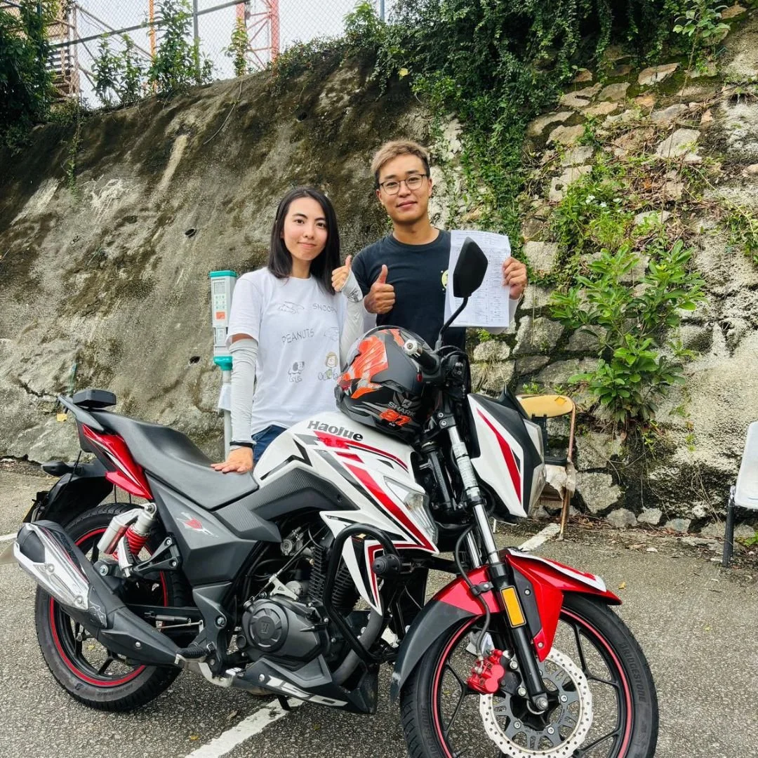 A man and woman standing beside a black and red Honda motorcycle outdoors, posing for the photo, with a rock wall and some greenery in the background.