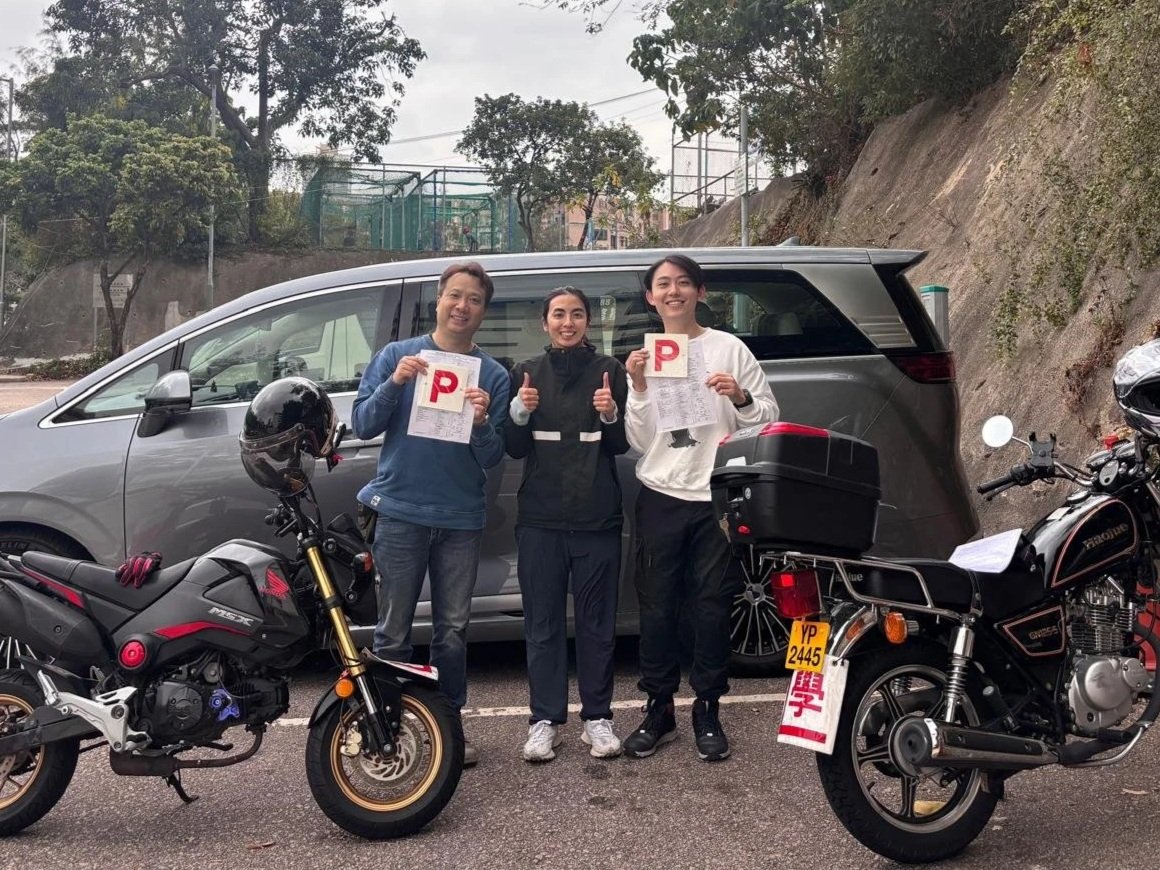 Three people standing in front of a silver van, holding driving license papers, with two motorcycles parked in front. The person on the left holds a large red 'P' sign, the person in the middle shows a thumbs-up, and the person on the right holds the