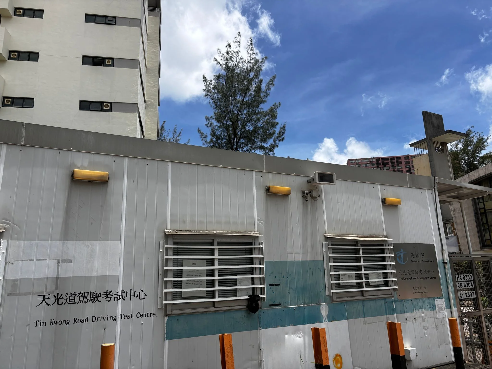 A sign in front of the Tin Kwong Road Driving Test Centre with Chinese and English text, a small barrier, and a blue sky with some clouds in the background.