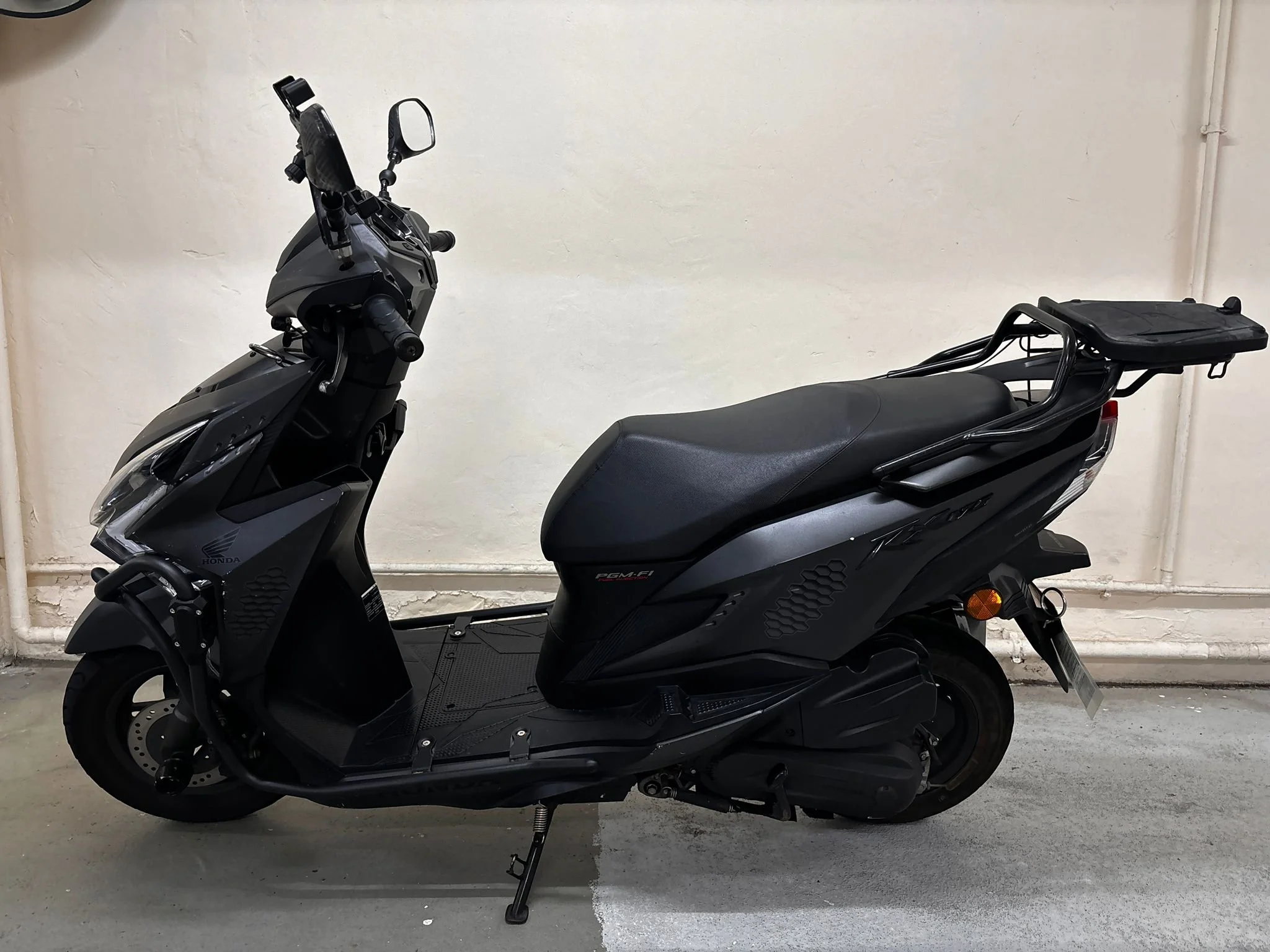 A black and gray Honda scooter parked indoors against a plain off-white wall, with a small rear cargo rack and a kickstand.