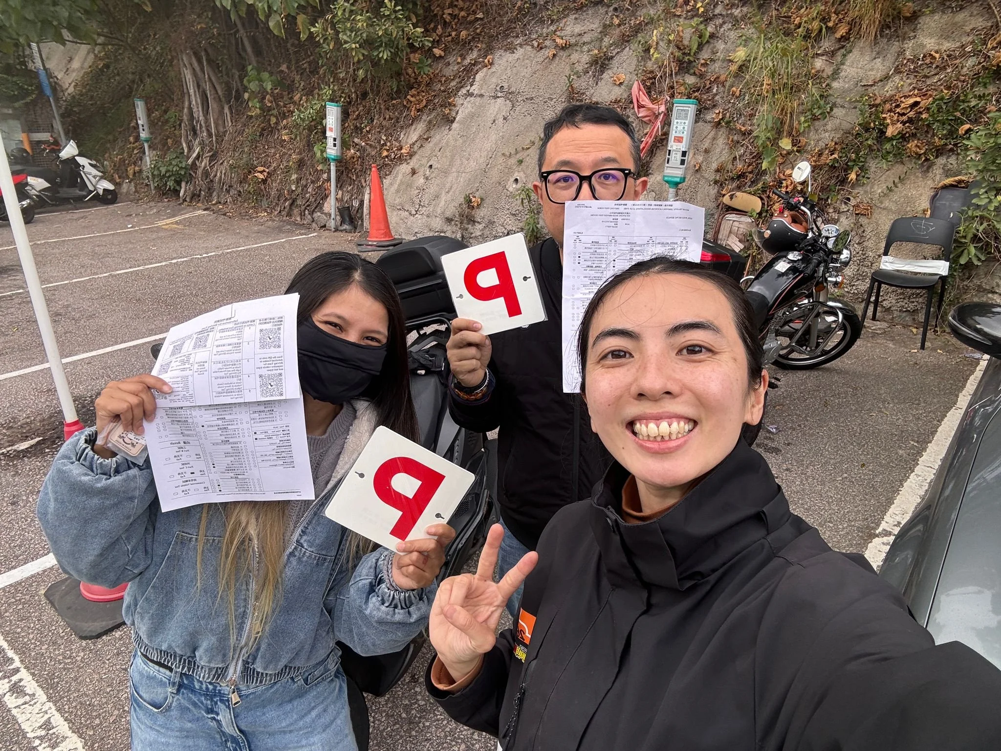 A group of three people taking a selfie at a parking lot, holding papers and protest signs, with motorcycles and parking meters in the background.