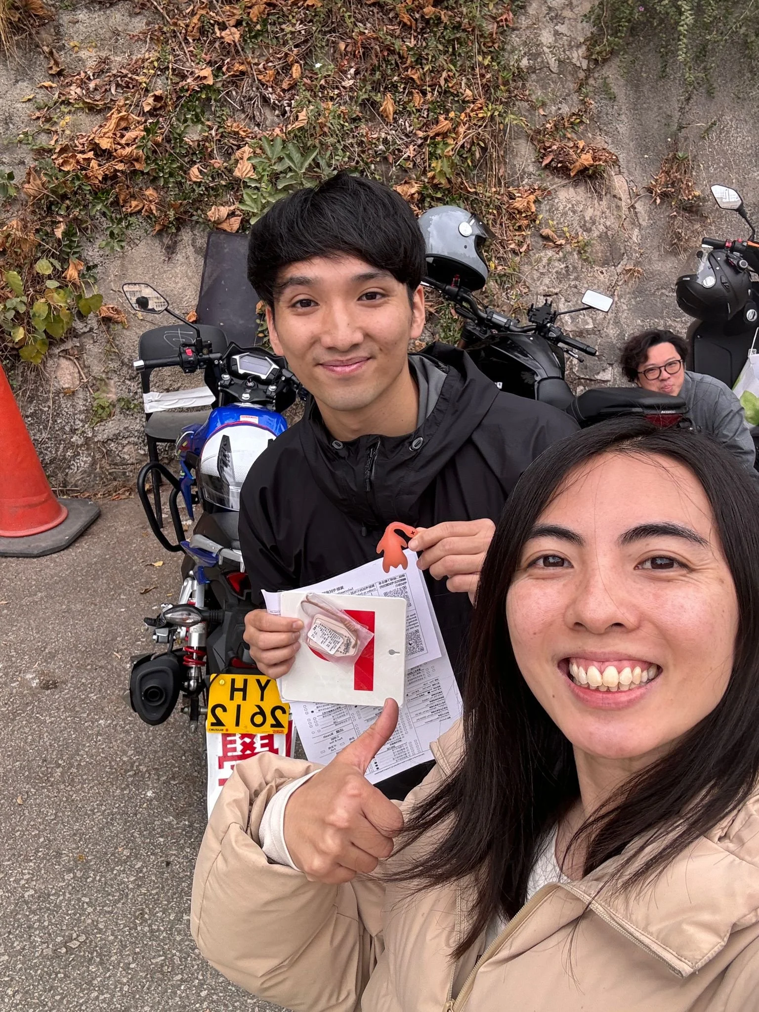Two women and a man taking a selfie outdoors. The man is smiling and holding a paper and a small orange object. The woman in front is smiling widely, and the woman in the background is sitting on a motorbike. The scene has fallen leaves and a concret