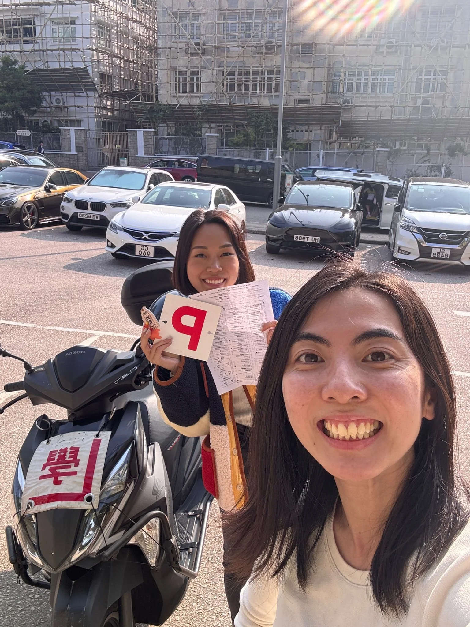 Two women smiling for a selfie outdoors, with parked cars and a building under construction with scaffolding in the background.