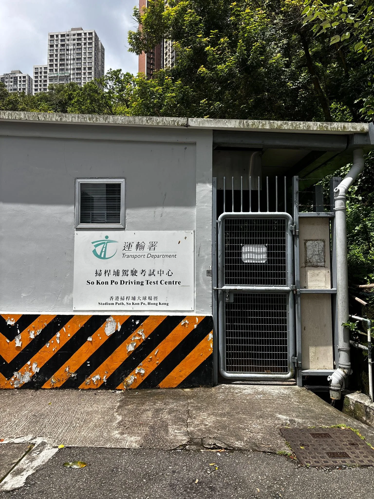 A small building with a sign that reads 'Transport Department' in English and Chinese, featuring a logo. The building has a window, a gated entrance, and a drainpipe on the right. The foreground shows a wet, textured asphalt surface with some fallen leaves, and in the background, tall modern buildings and green trees under a partly cloudy sky.