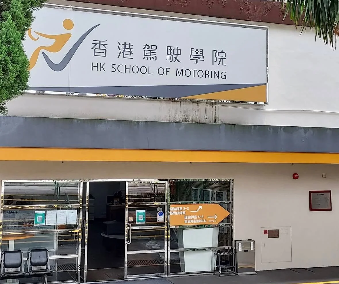 Sign for HK School of Motoring with logo, Chinese characters, and English name, located outside a building with glass doors and seating area.