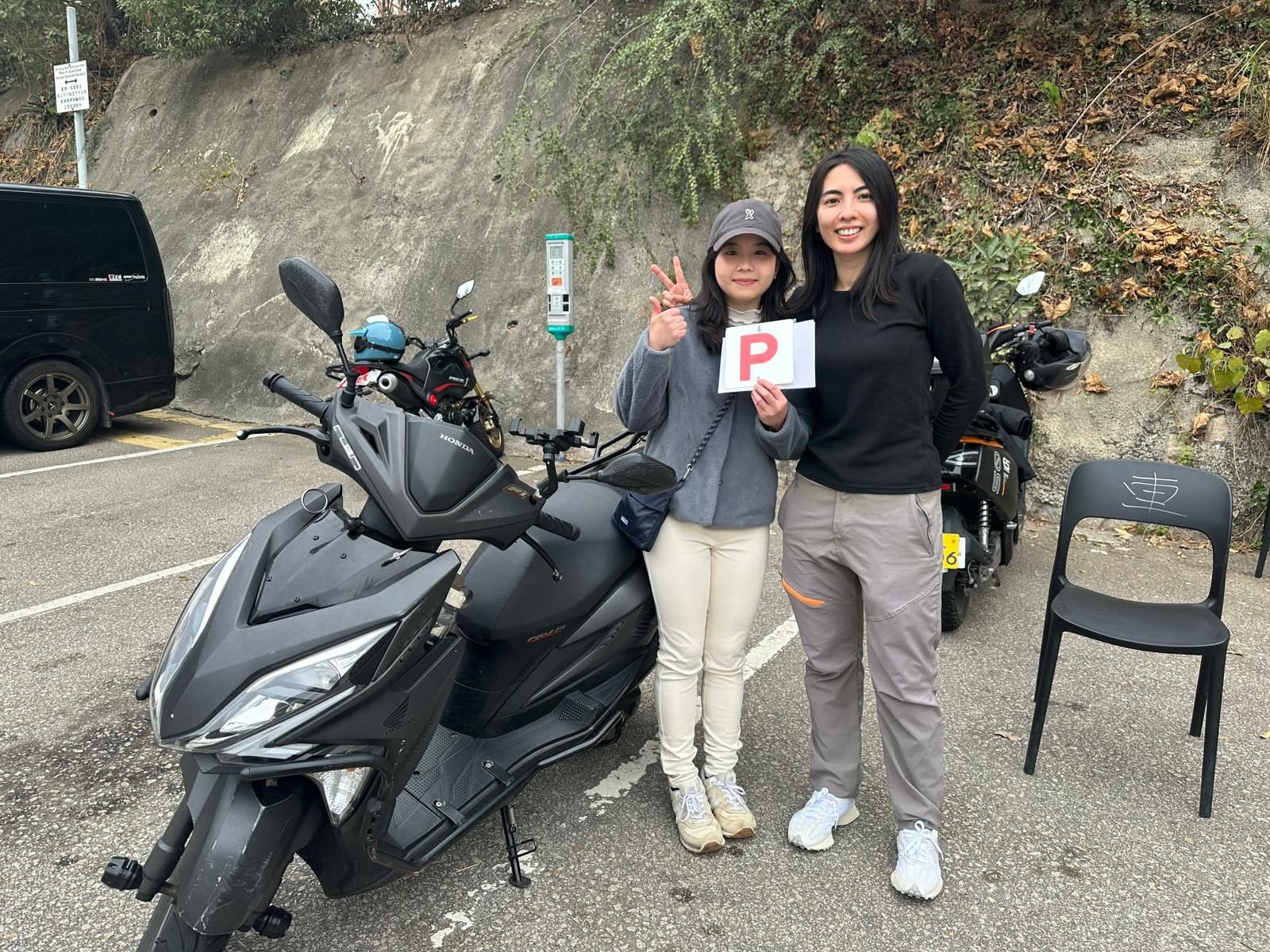 Two women standing in a parking lot beside a scooter and a motorcycle, with a chair nearby. One woman is holding a sign with a red letter 'P'. The woman on the left is making a peace sign with her fingers, and both are smiling. Background includes ca