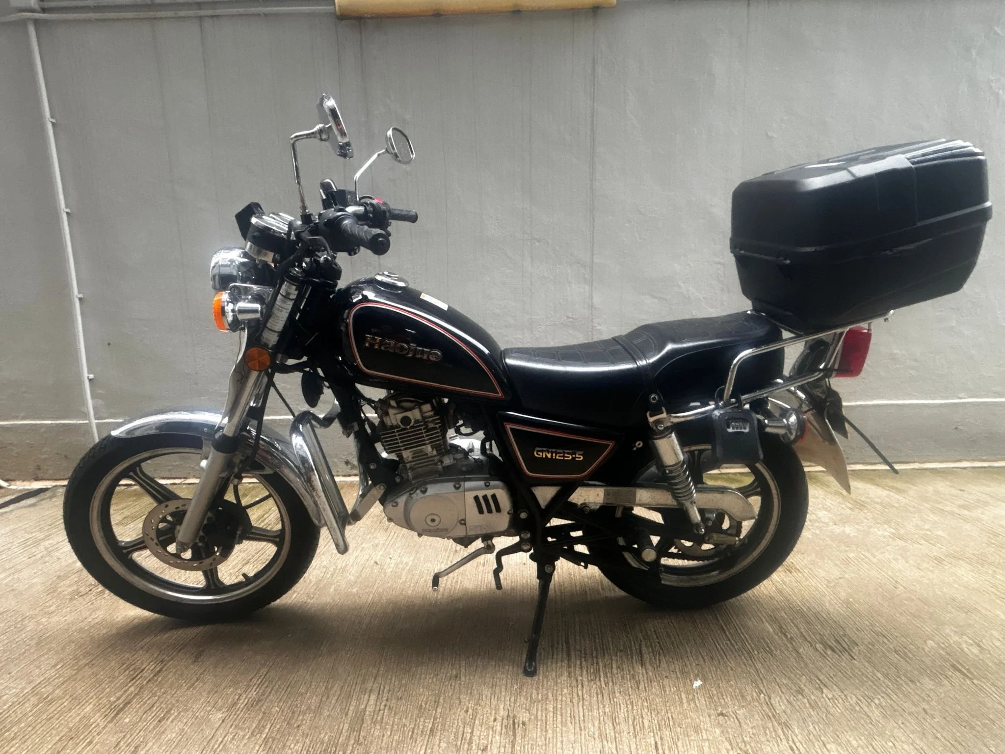 Black Honda motorcycle with a top case, parked indoors against a light gray wall, on a concrete floor.