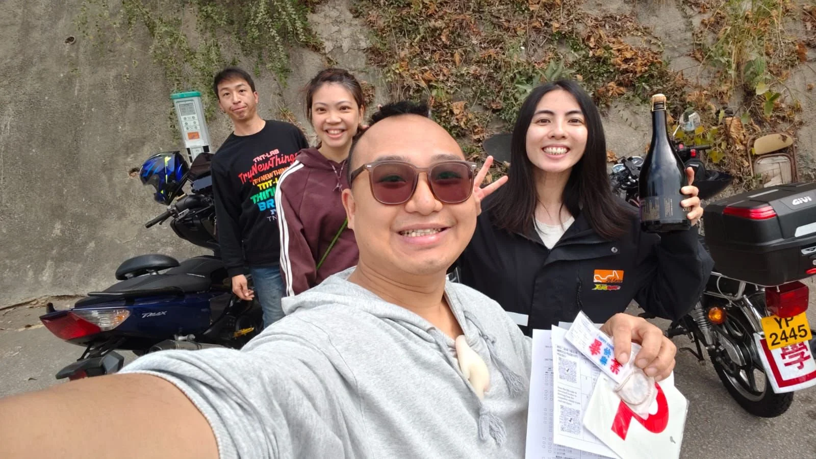Group of four friends taking a selfie outdoors with motorbikes and a rocky background.