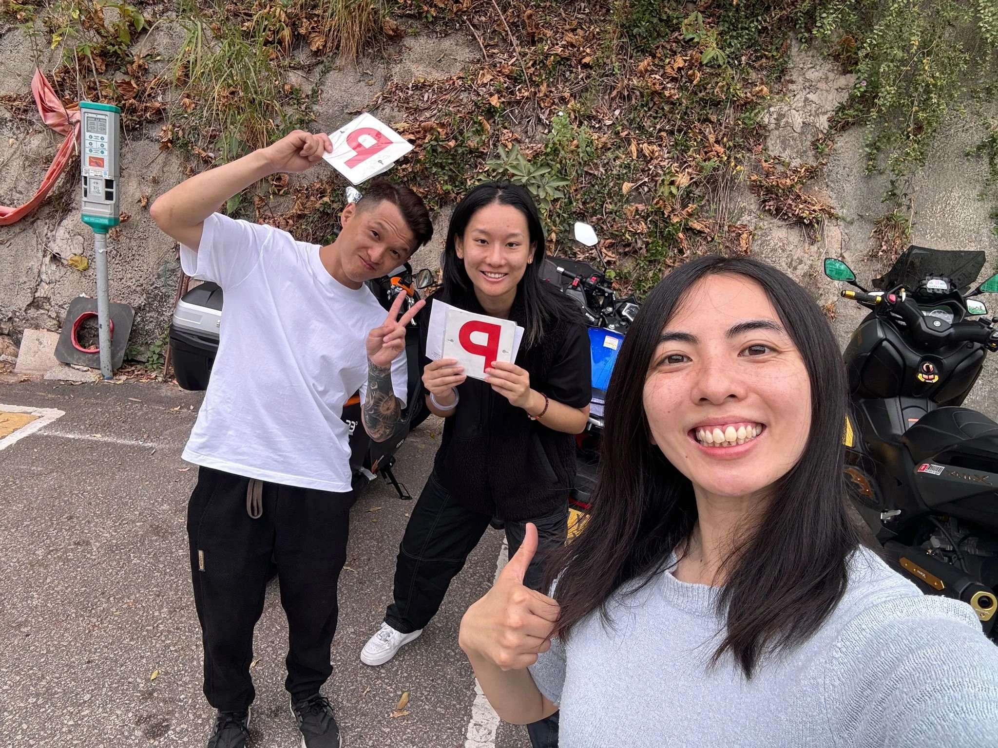 Group of three friends taking a selfie outdoors, holding pizza boxes, with motorcycles and a stone wall covered with plants in the background.