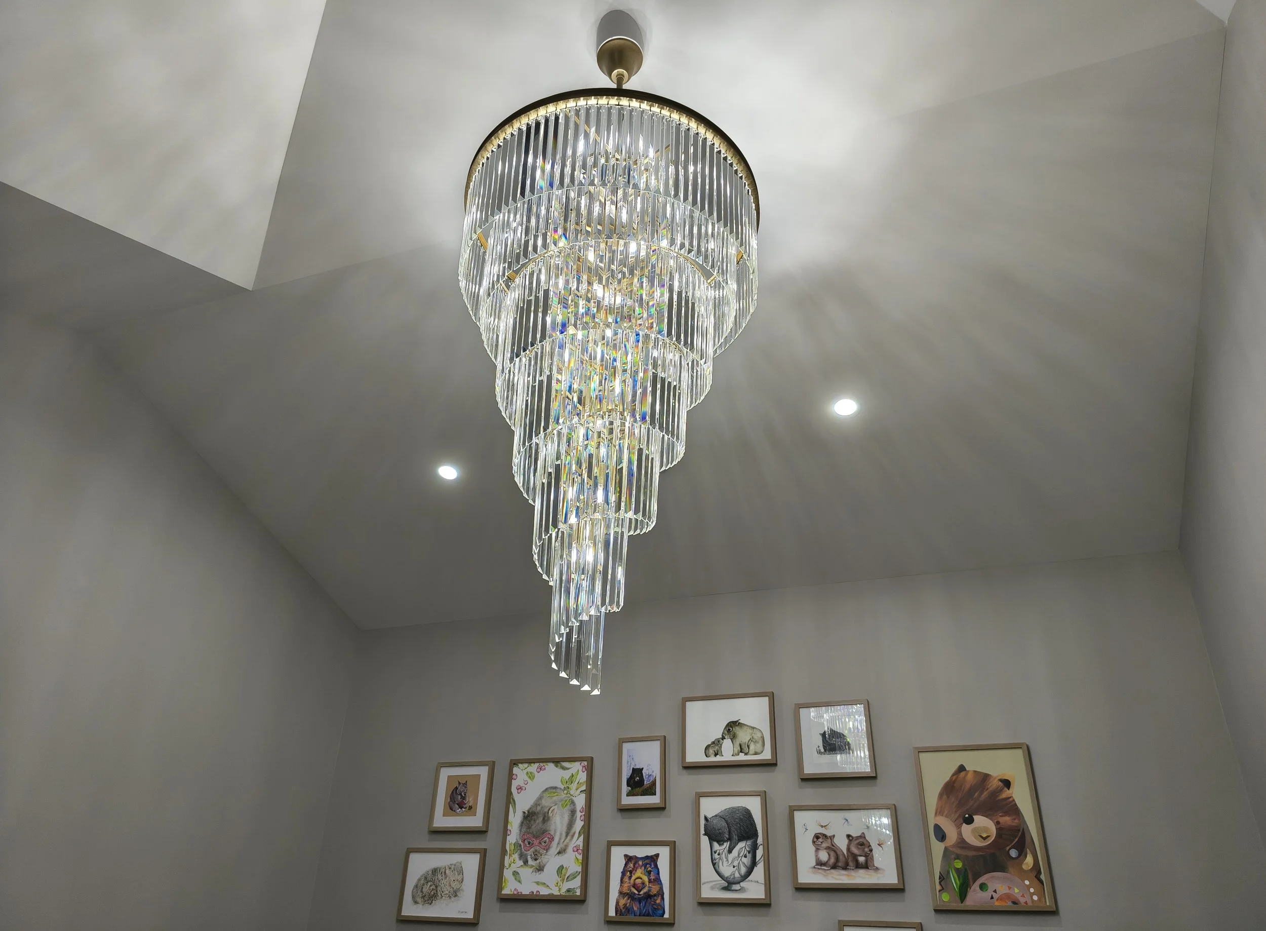 A large, multi-tiered crystal chandelier hanging from a ceiling with two small recessed lights, above a gallery wall with various framed animal artwork.