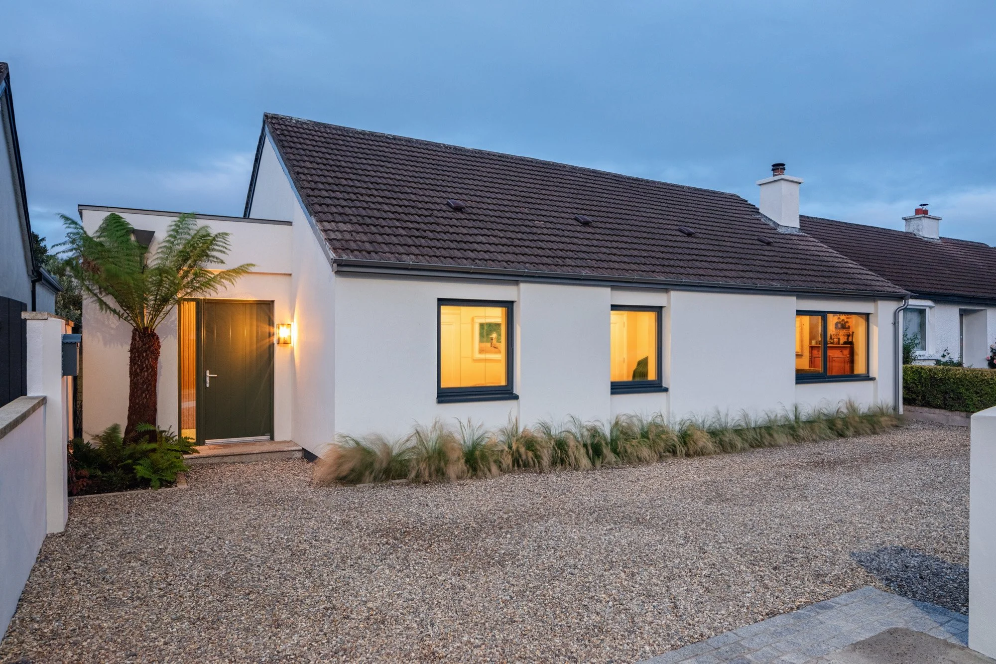 Front exterior of a modern house with white walls, a dark green front door, and three illuminated windows, surrounded by a gravel driveway and landscaping with a palm tree and ornamental grasses, during dusk.