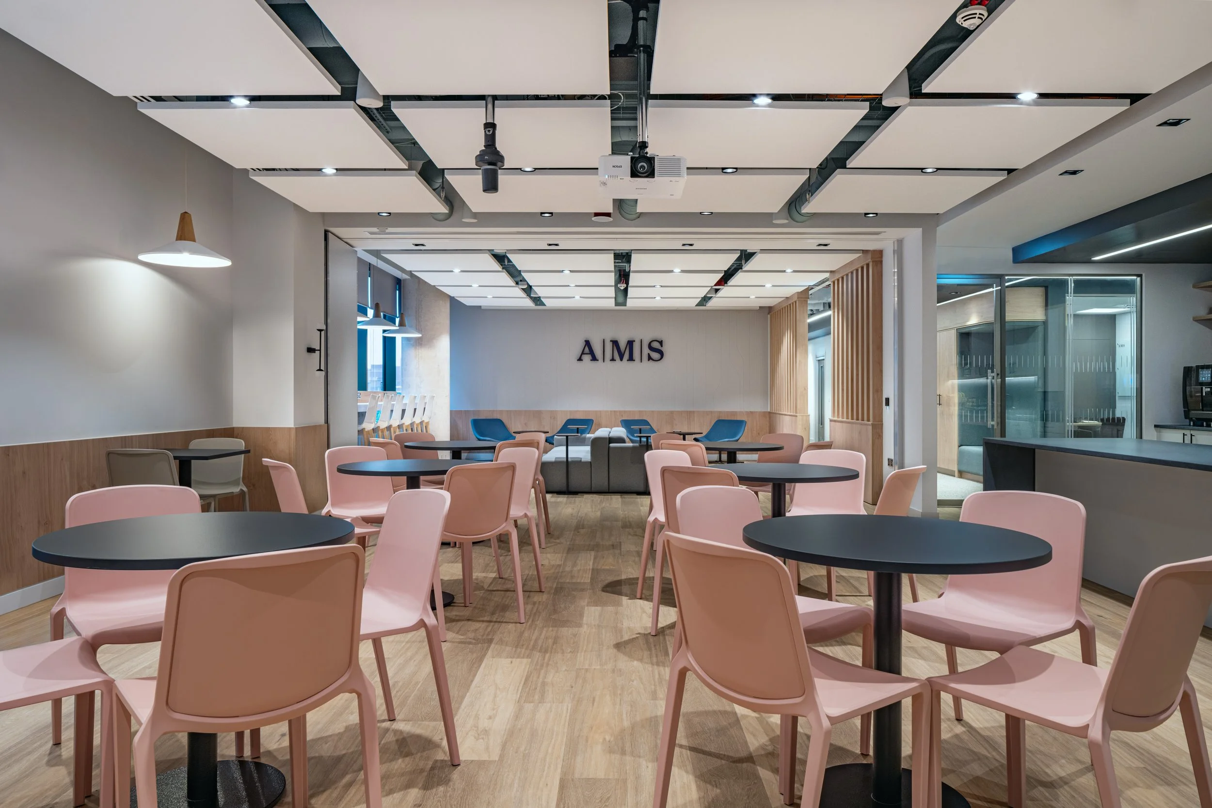Modern cafe with pink chairs, black tables, and a wooden floor. The wall at the back has the letters 'A M S' on it, with blue chairs and a sofa in the seating area.