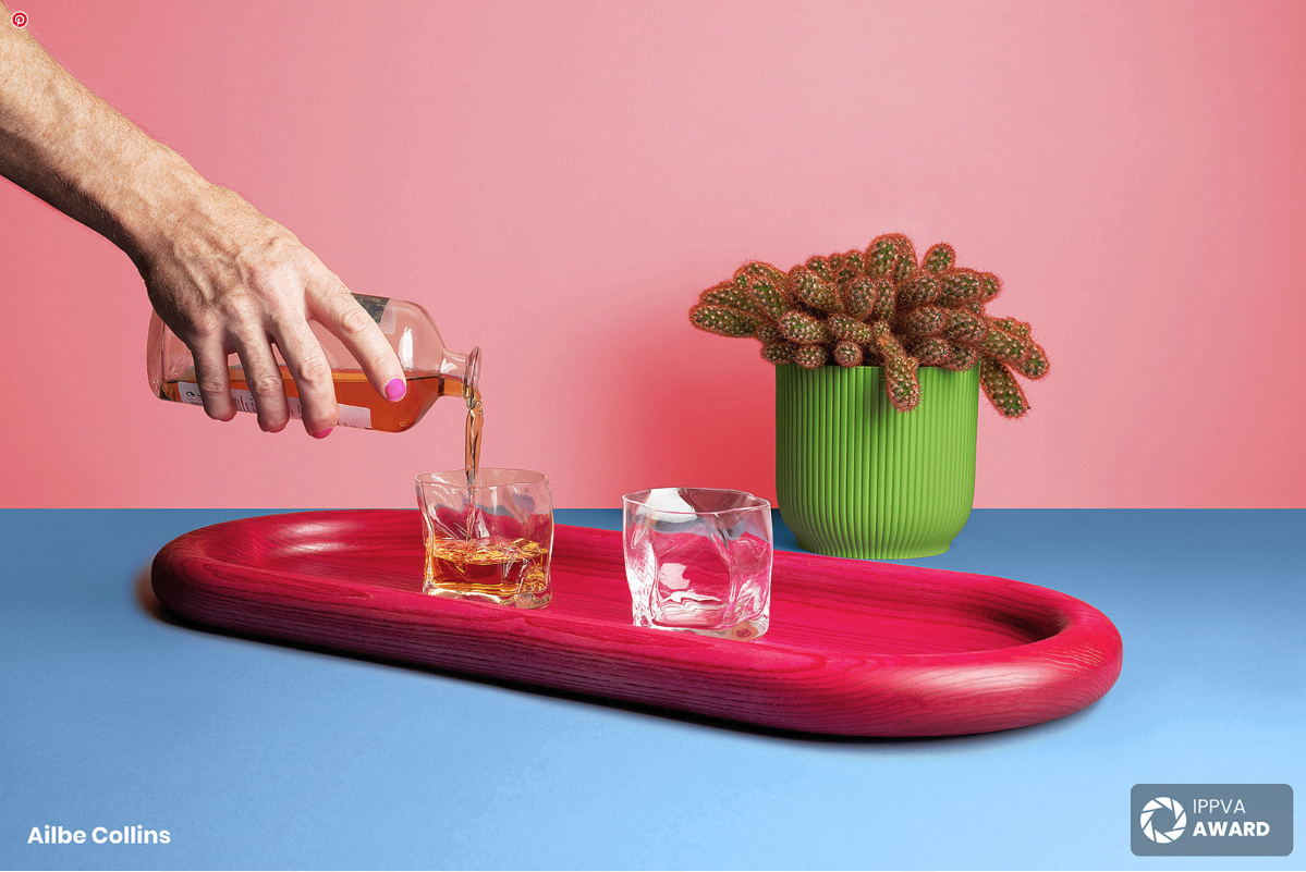 A hand with pink painted nails pouring a drink into a glass on a pink wooden tray, with two empty glasses and a green potted cactus plant in a green ribbed pot on a blue surface against a pink background.